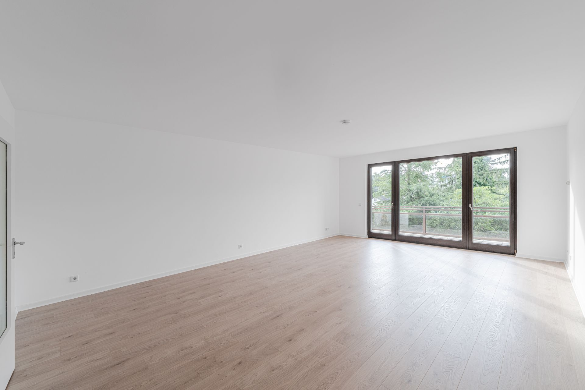 Helles, leeres Zimmer mit Holzfußboden und großer Fensterfront mit Blick ins Grüne.