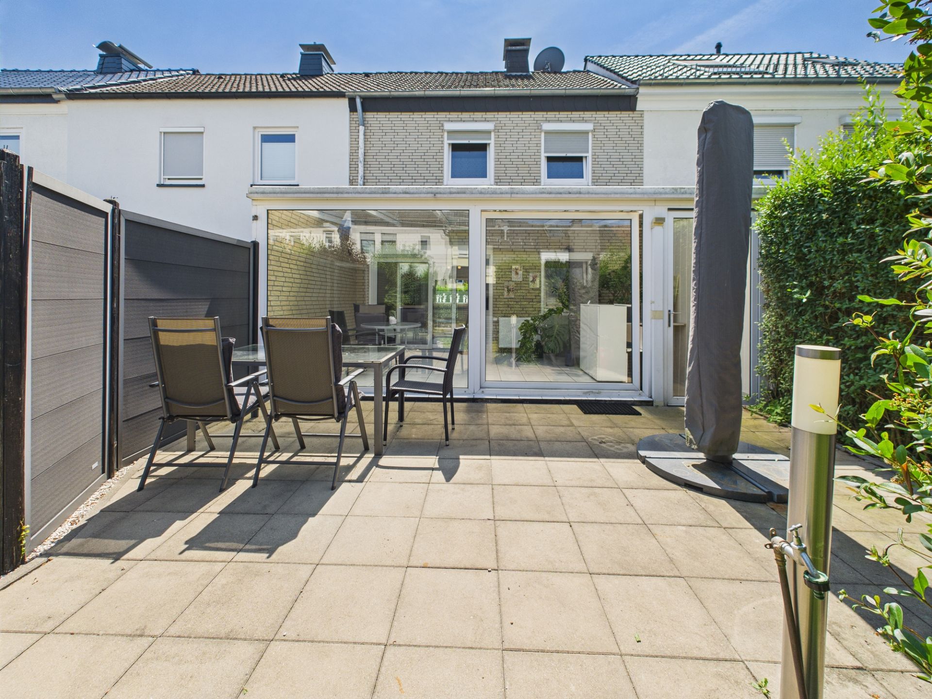 Gepflasterte Terrasse mit Tisch, vier Stühlen und großem Sonnenschirm, angrenzend an verglastes Haus.