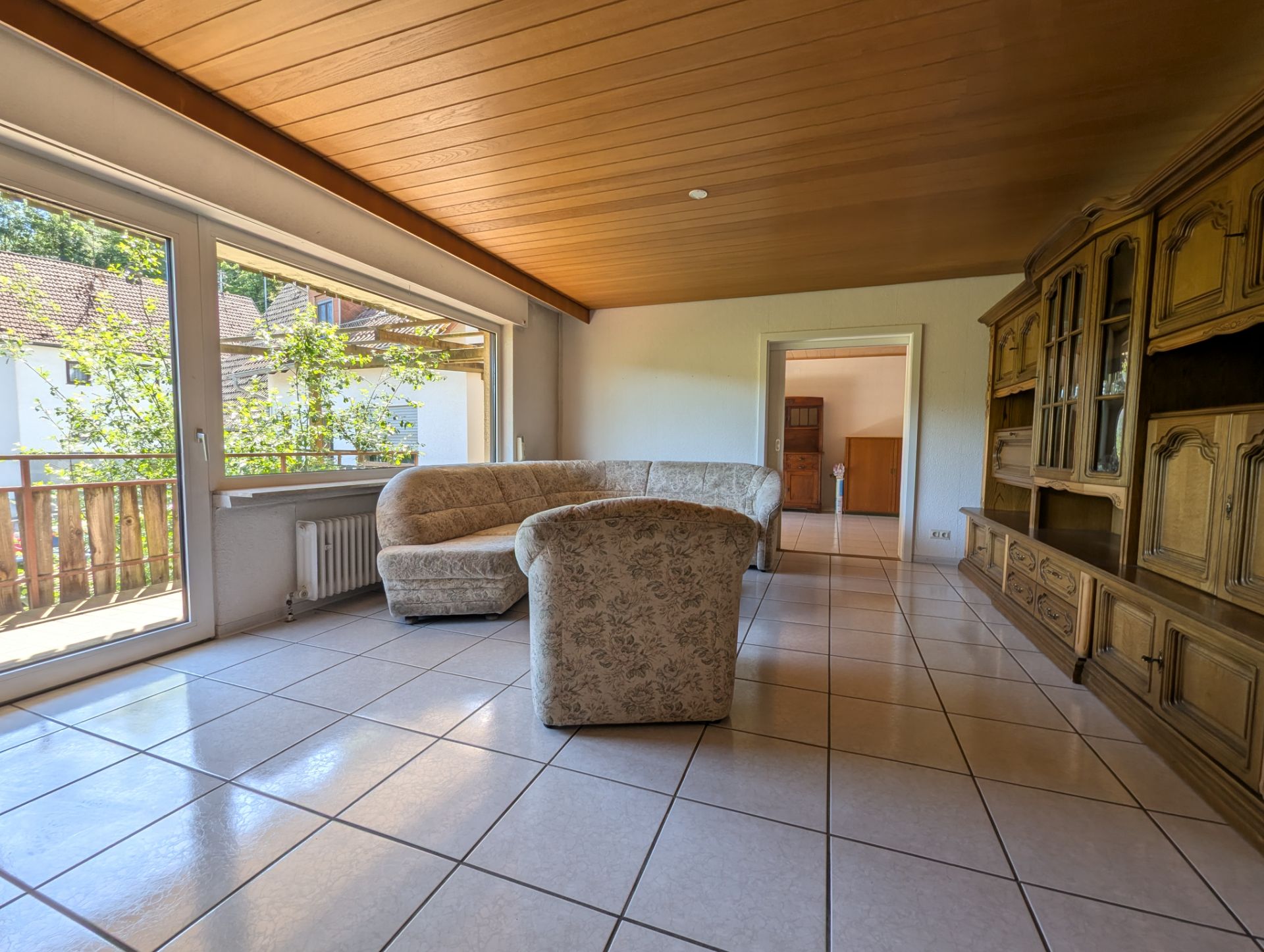 Wohnzimmer mit großer Fensterfront, Ecksofa, Sessel und Holzschrank auf Fliesenboden.