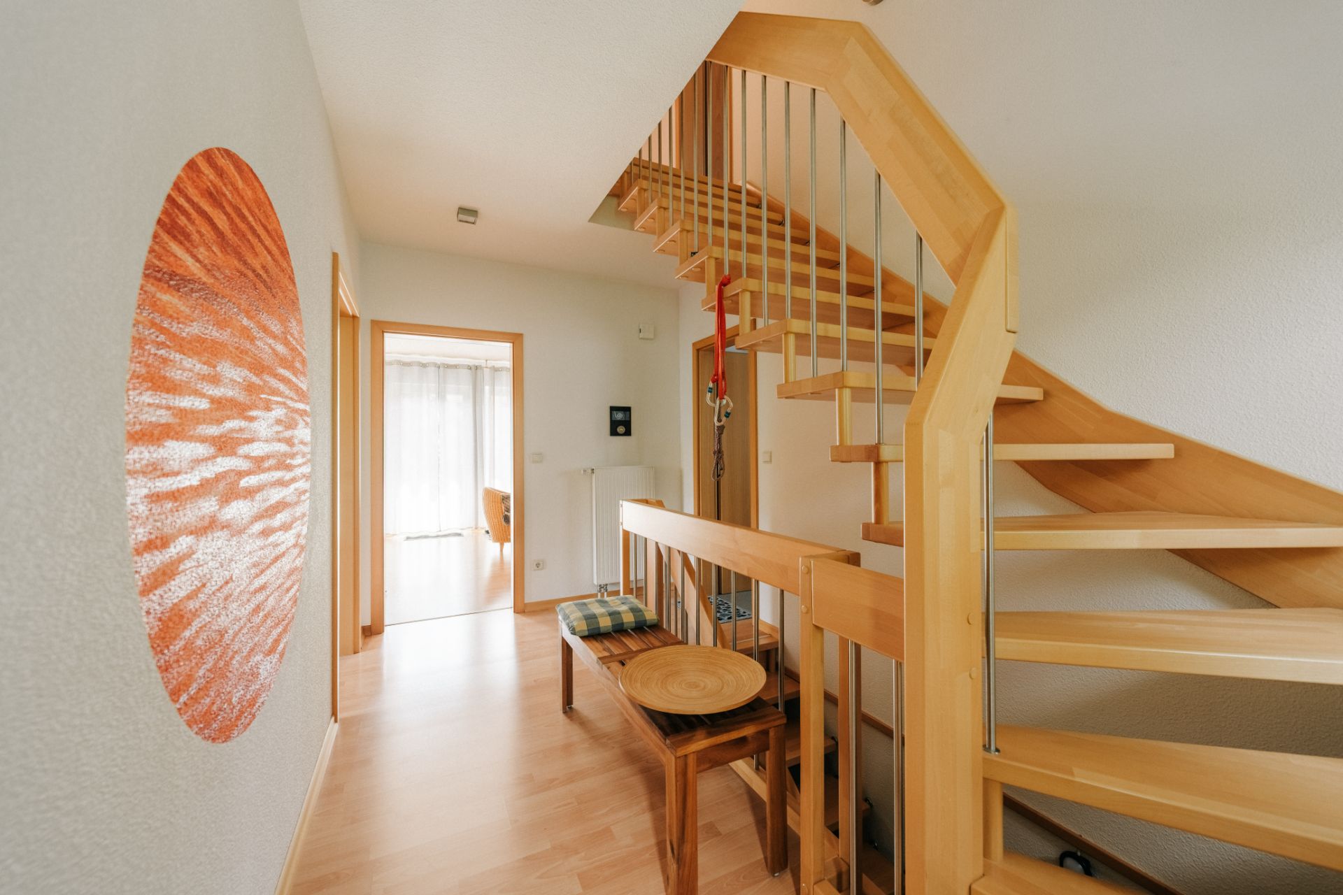 Heller Flur mit Holztreppe, Sitzbank und rundem Wandbild.