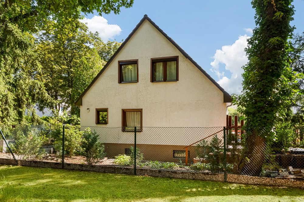 Weißes Einfamilienhaus mit Spitzdach, braunen Fenstern und Garten, umgeben von einem Zaun.