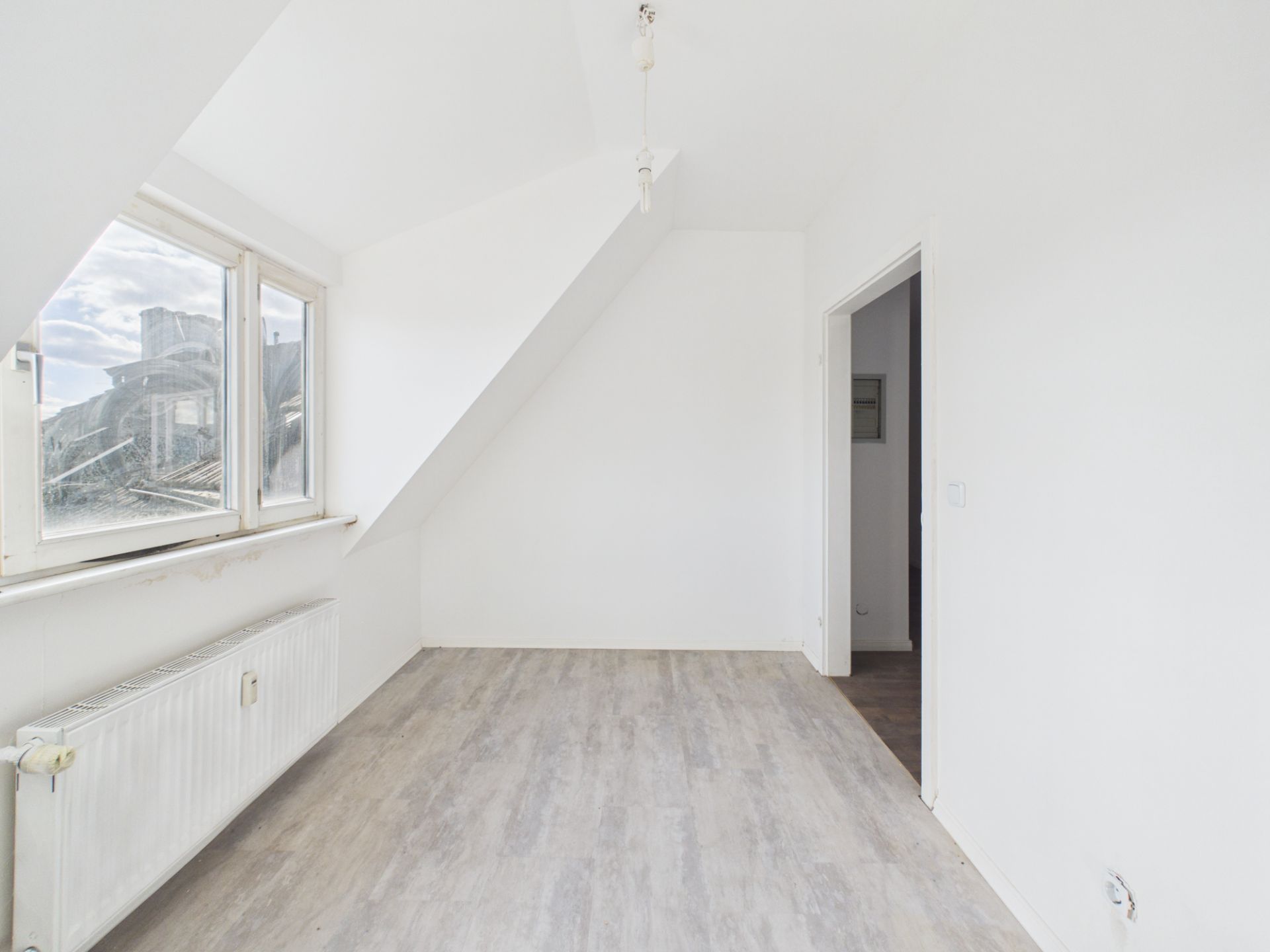 Helles, leeres Zimmer mit Dachschräge, Fenster, Heizkörper und Holzboden.