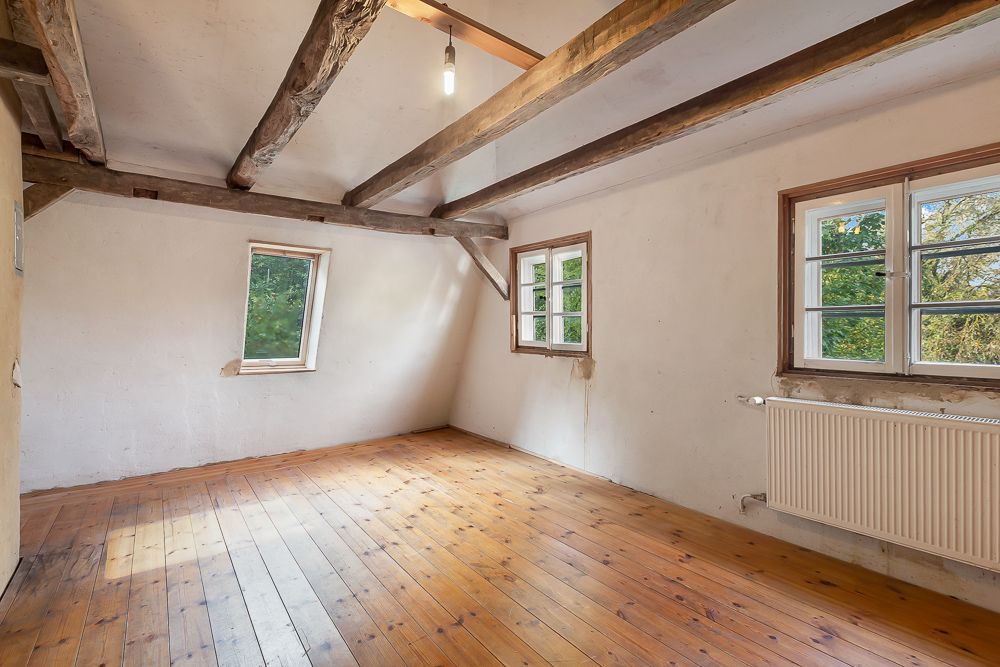Leeres Zimmer mit Holzboden, sichtbaren Holzdeckenbalken und zwei Fenstern.