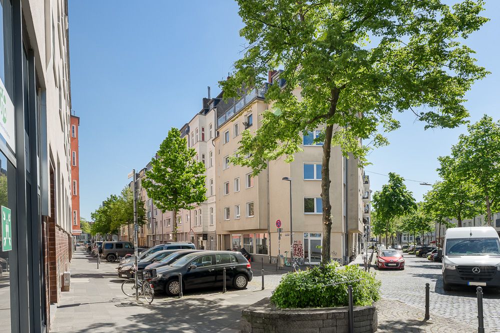 Wohnstraße mit geparkten Autos, Bäumen und mehrstöckigen Wohnhäusern bei sonnigem Wetter.