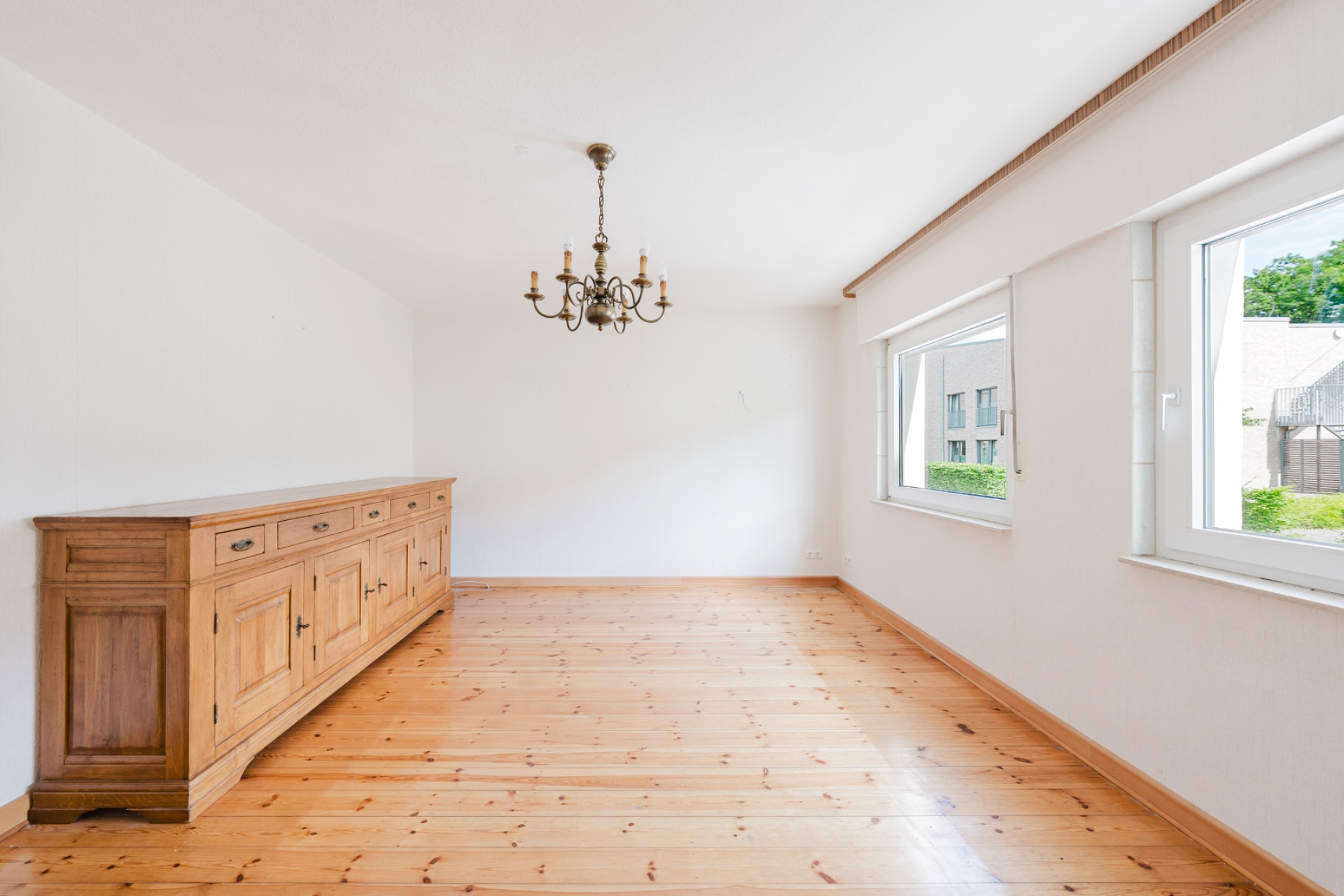 Leerer Raum mit Holzfußboden, Kronleuchter, Sideboard und zwei Fenstern.