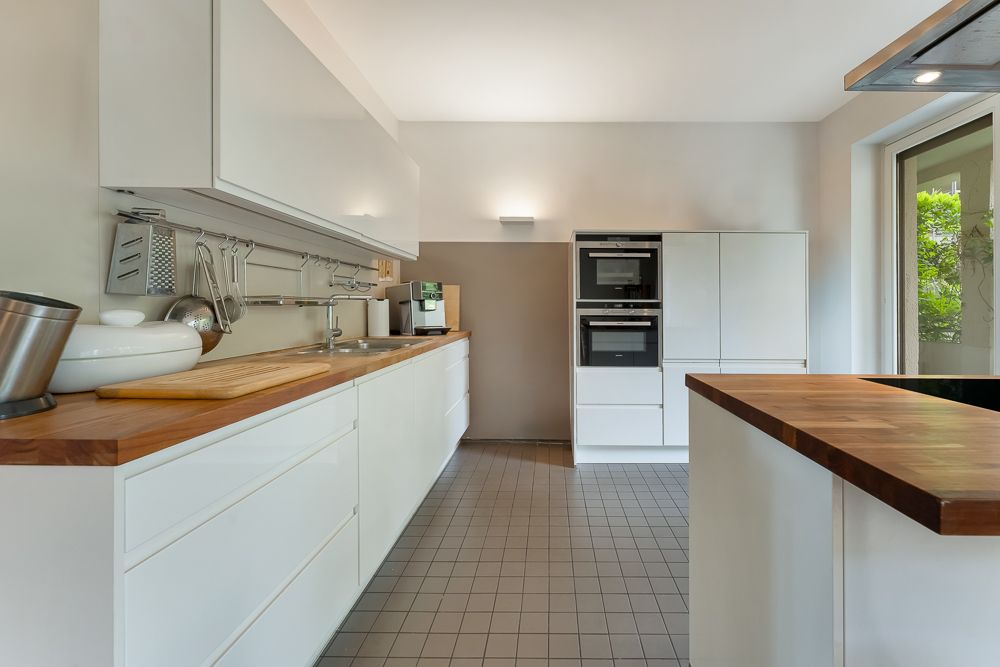 Moderne Küche mit weißen Schränken, Holzar­beitsplatten und großen Fenstern.
