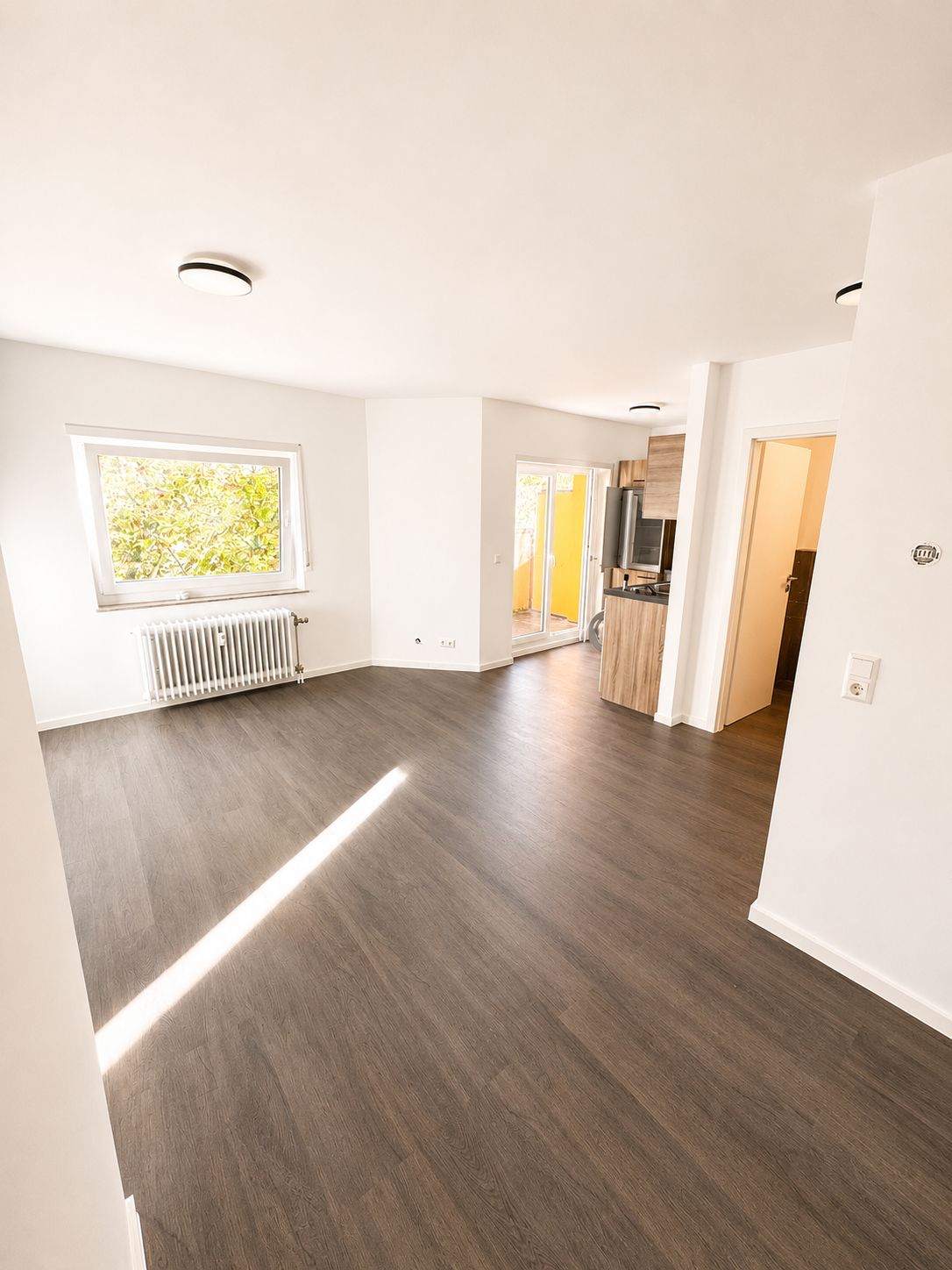 Helles Wohnzimmer mit großem Fenster, offen zur Küche, mit braunem Boden.