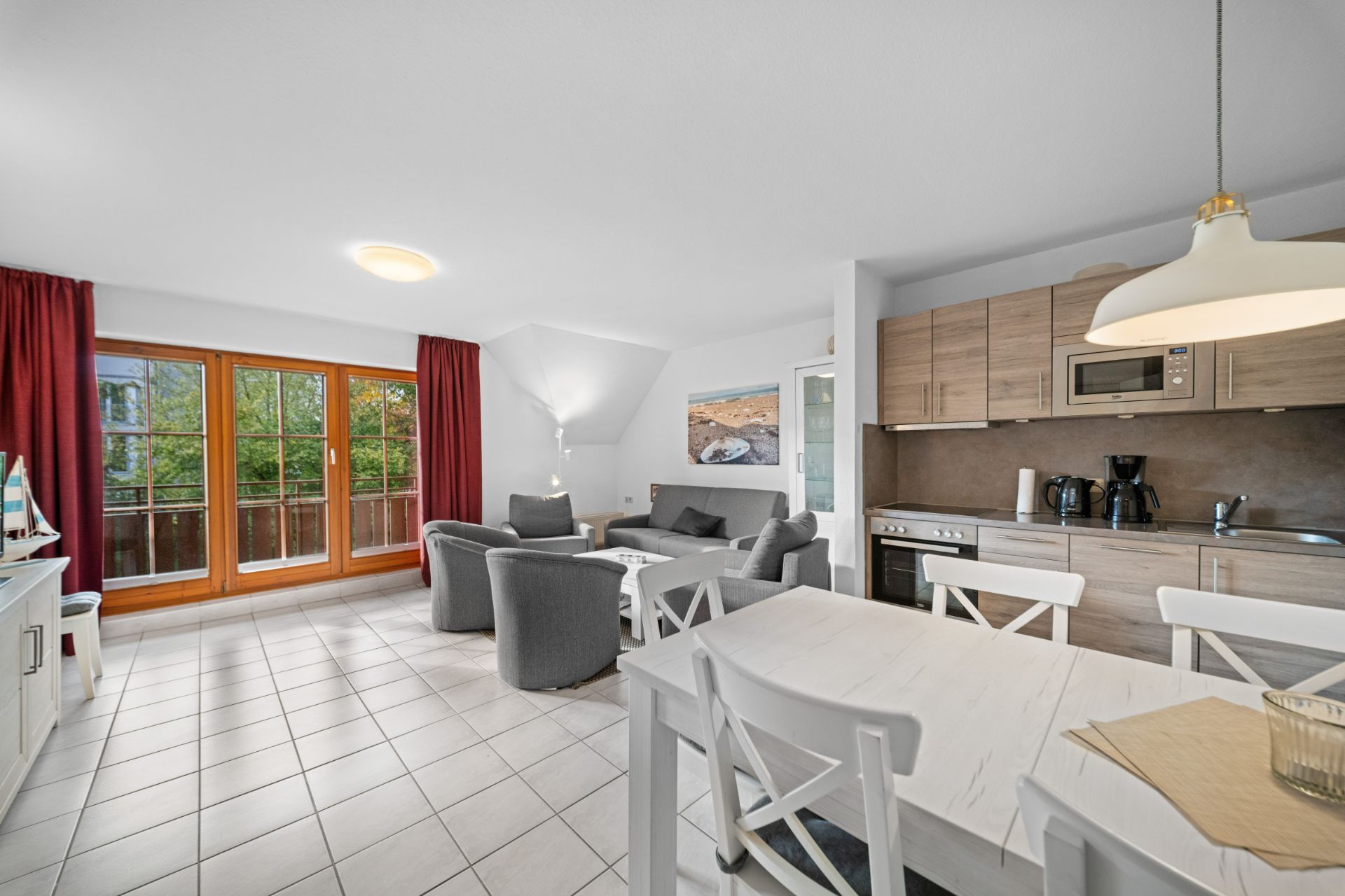 Helles Wohnzimmer mit offener Küche, Esstisch, Sofa und großen Fenstern mit roten Vorhängen.