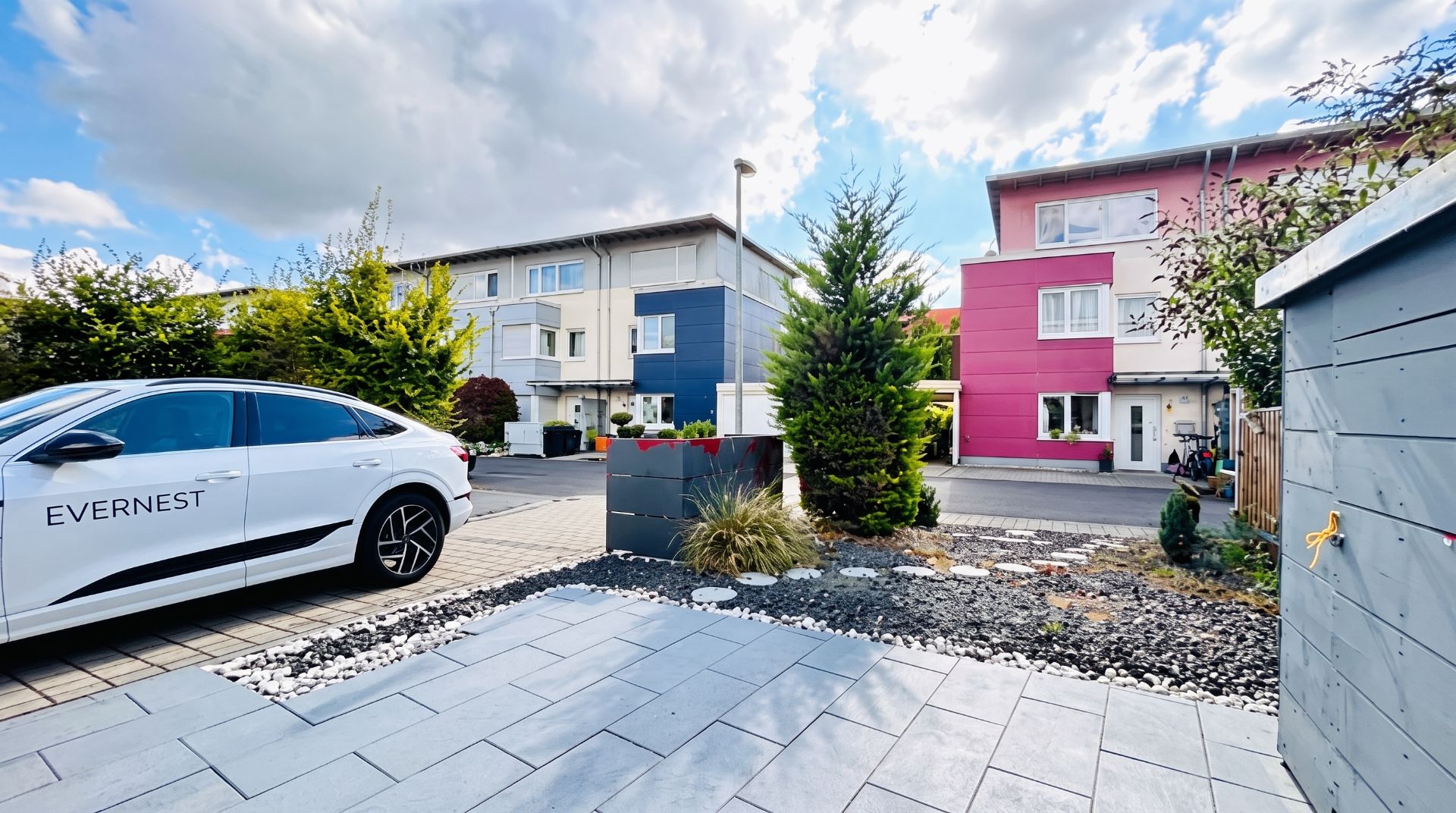 Moderner Vorgarten mit Steinplatten, Einfahrt, Auto und bunten Mehrfamilienhäusern im Hintergrund.