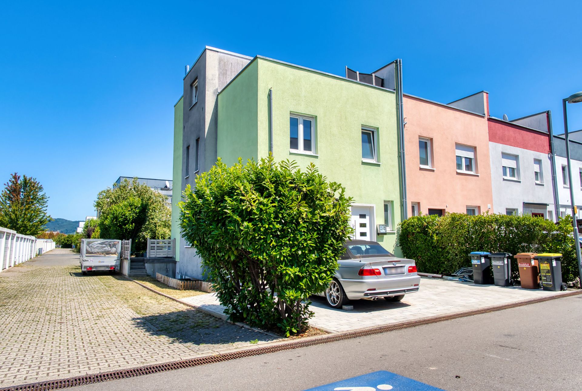 Moderne Reihenhäuser mit bunten Fassaden, Auto und grünen Büschen vor dem Haus, sonniger Tag.