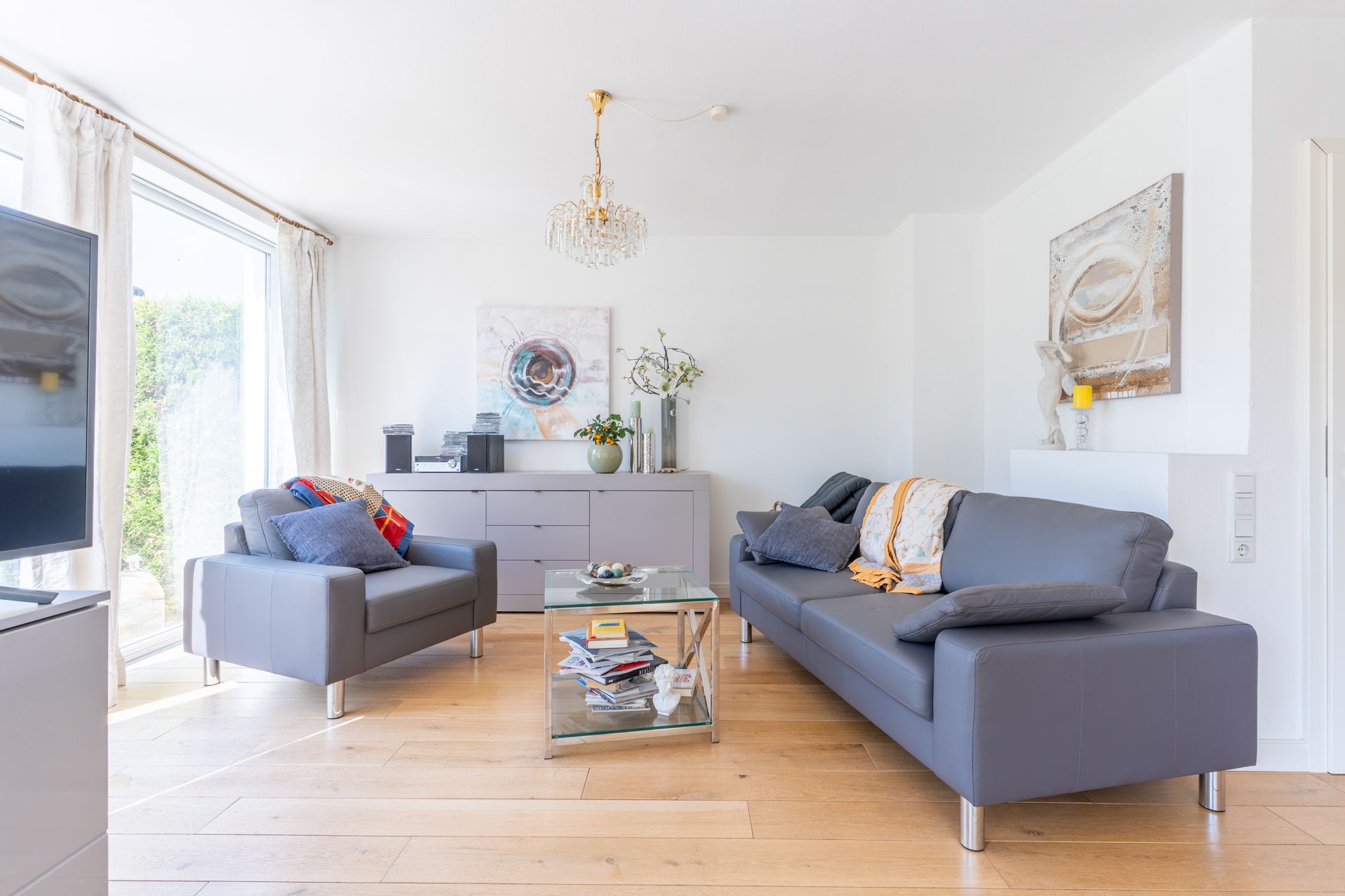 Helles Wohnzimmer mit grauem Sofa, Sessel, Glastisch und Kommode vor großen Fenstern.
