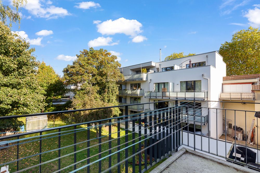 Blick von einem Balkon auf ein modernes Wohnhaus und einen Garten mit Bäumen.