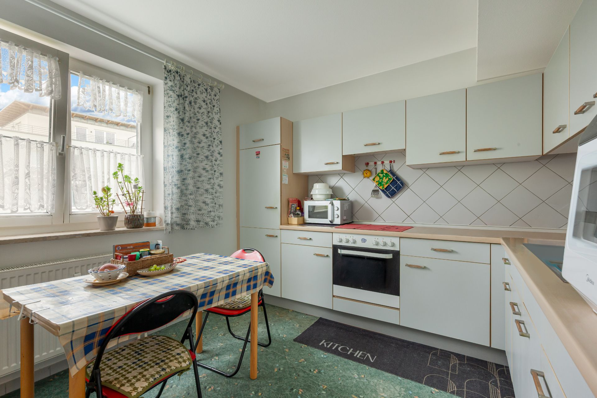 Helle Küche mit Esstisch, Stühlen, weißen Schränken und Herd, Fenster mit Pflanzen.