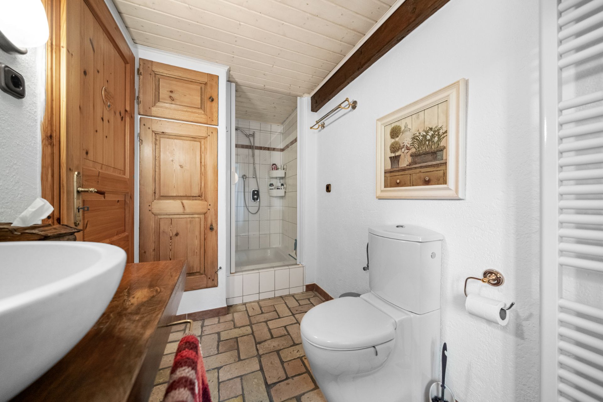 Helles Badezimmer mit Toilette, Waschbecken, Dusche, Holzschrank und einem Bild an der Wand.