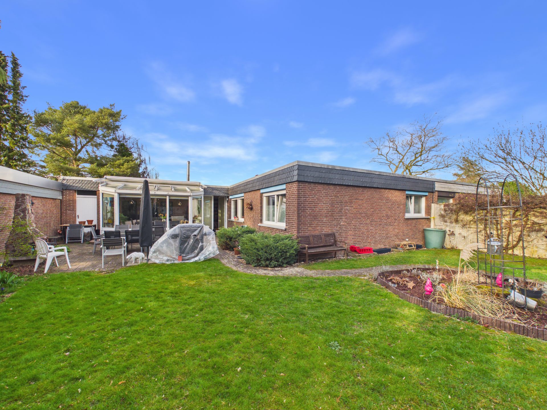 Backsteinbungalow mit Flachdach, Terrasse mit Gartenmöbeln und gepflegtem Garten im Vordergrund.