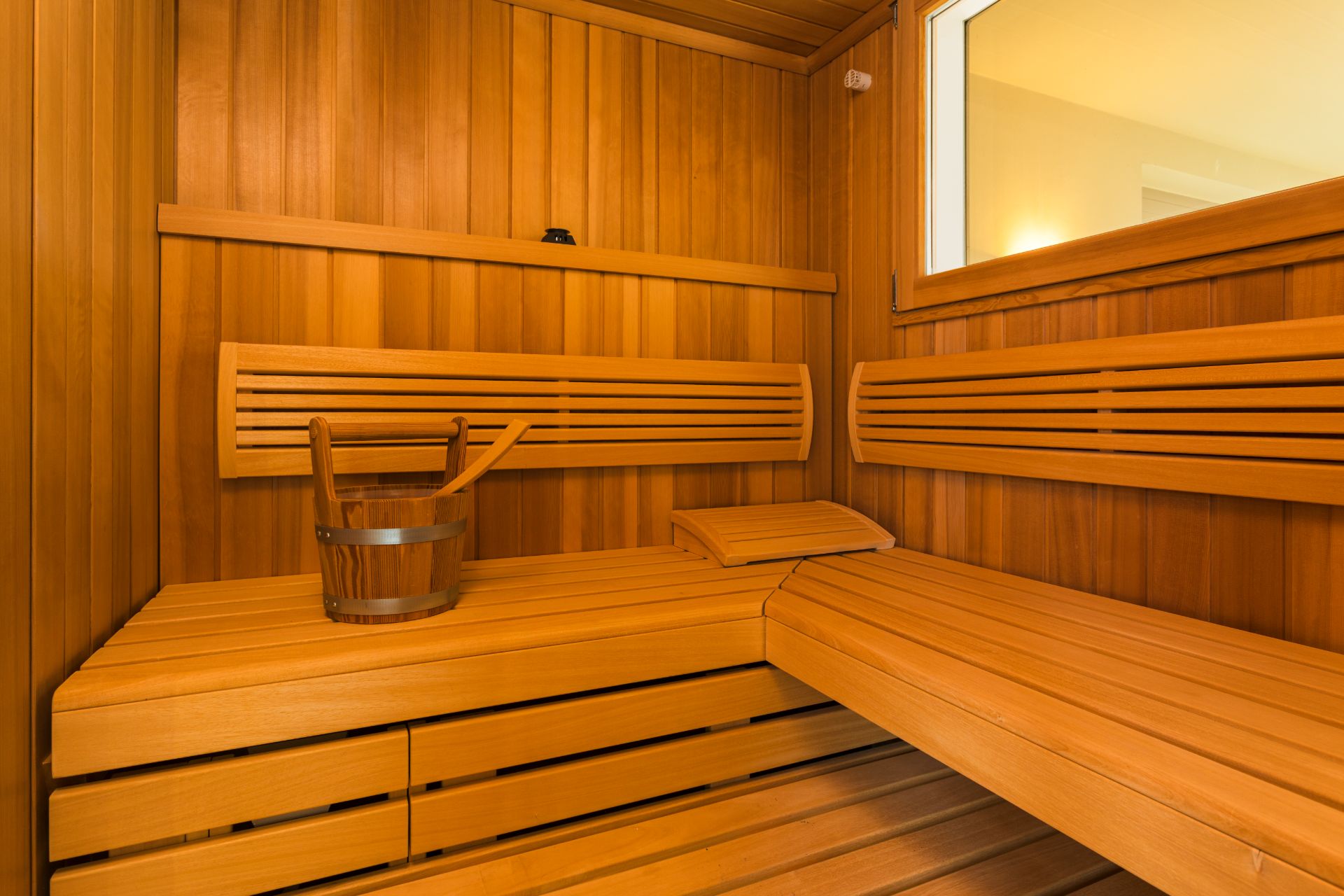 Innenansicht einer Holzsauna mit Bänken, Eimer und einem Fenster.