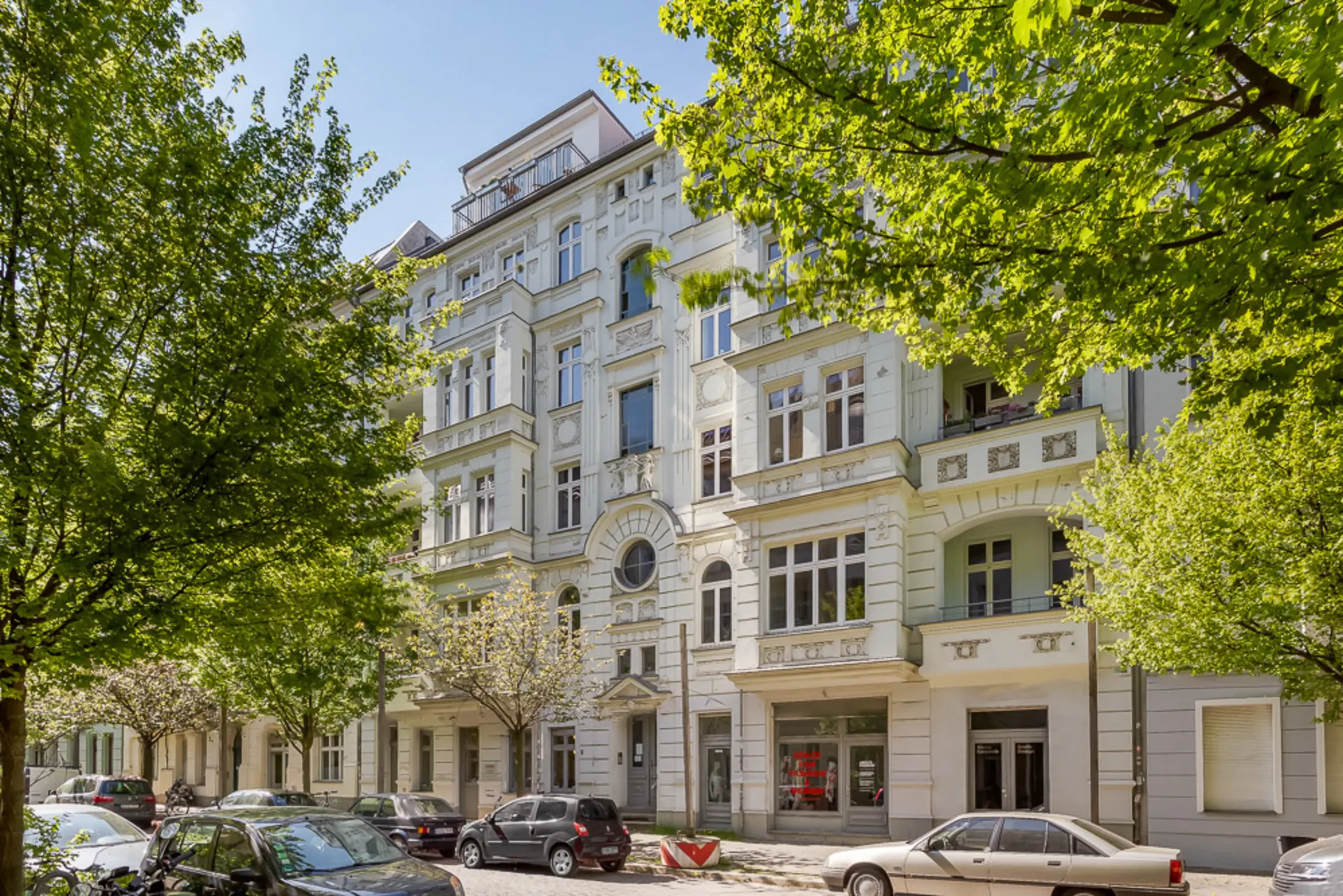 Mehrstöckiges Wohnhaus mit Balkonen und reicher Fassadendekoration, Autos und Bäume davor.