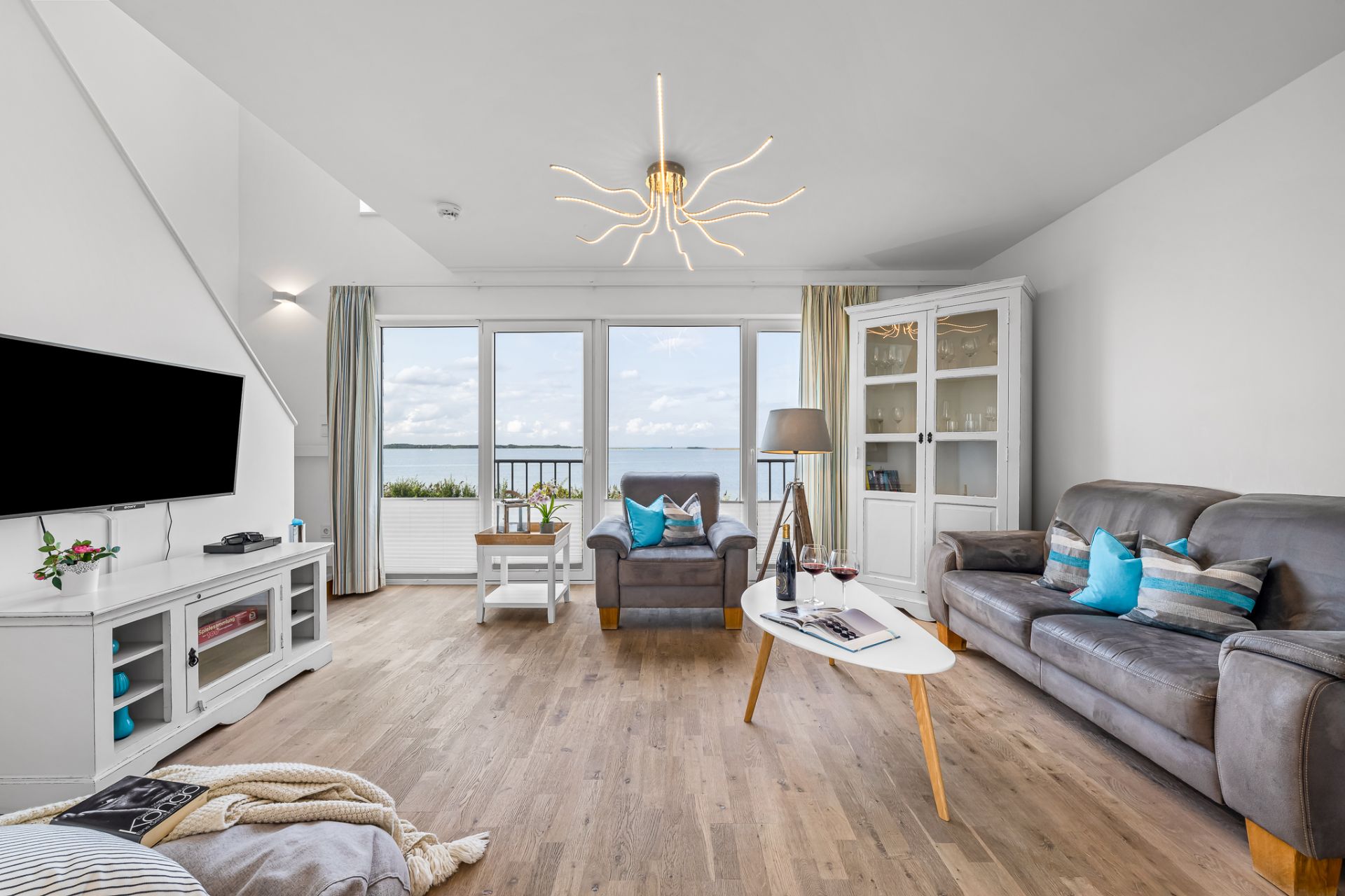 Helles Wohnzimmer mit Sofa, Sessel, Fernseher, Glastür und Blick auf das Meer.