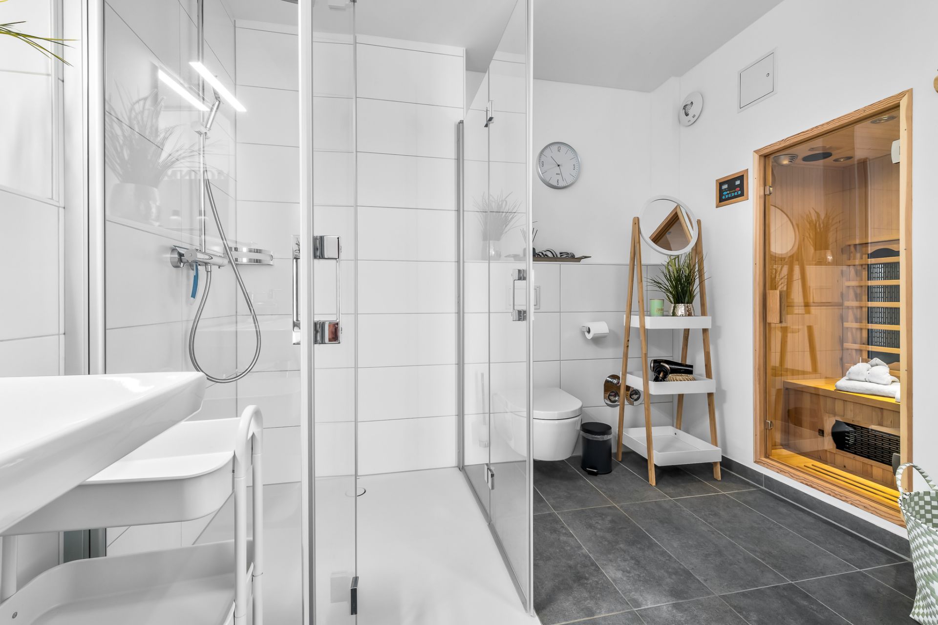 Modernes Badezimmer mit Glasdusche, Toilette, Regal und Holzsauna auf dunklem Fliesenboden.