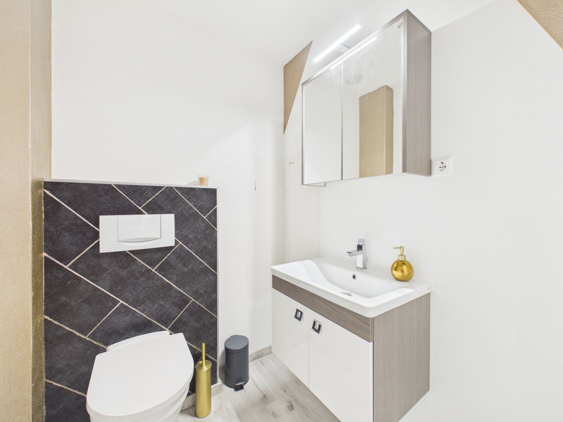 Modernes Badezimmer mit WC, Waschbecken, Spiegelschrank und goldfarbenen Accessoires.