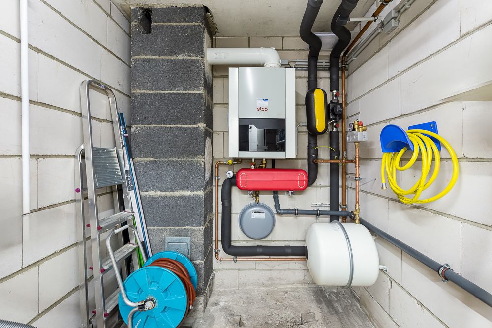 Technikraum mit Heizungsanlage, Rohren, Leiter und gelbem Schlauch an der Wand.