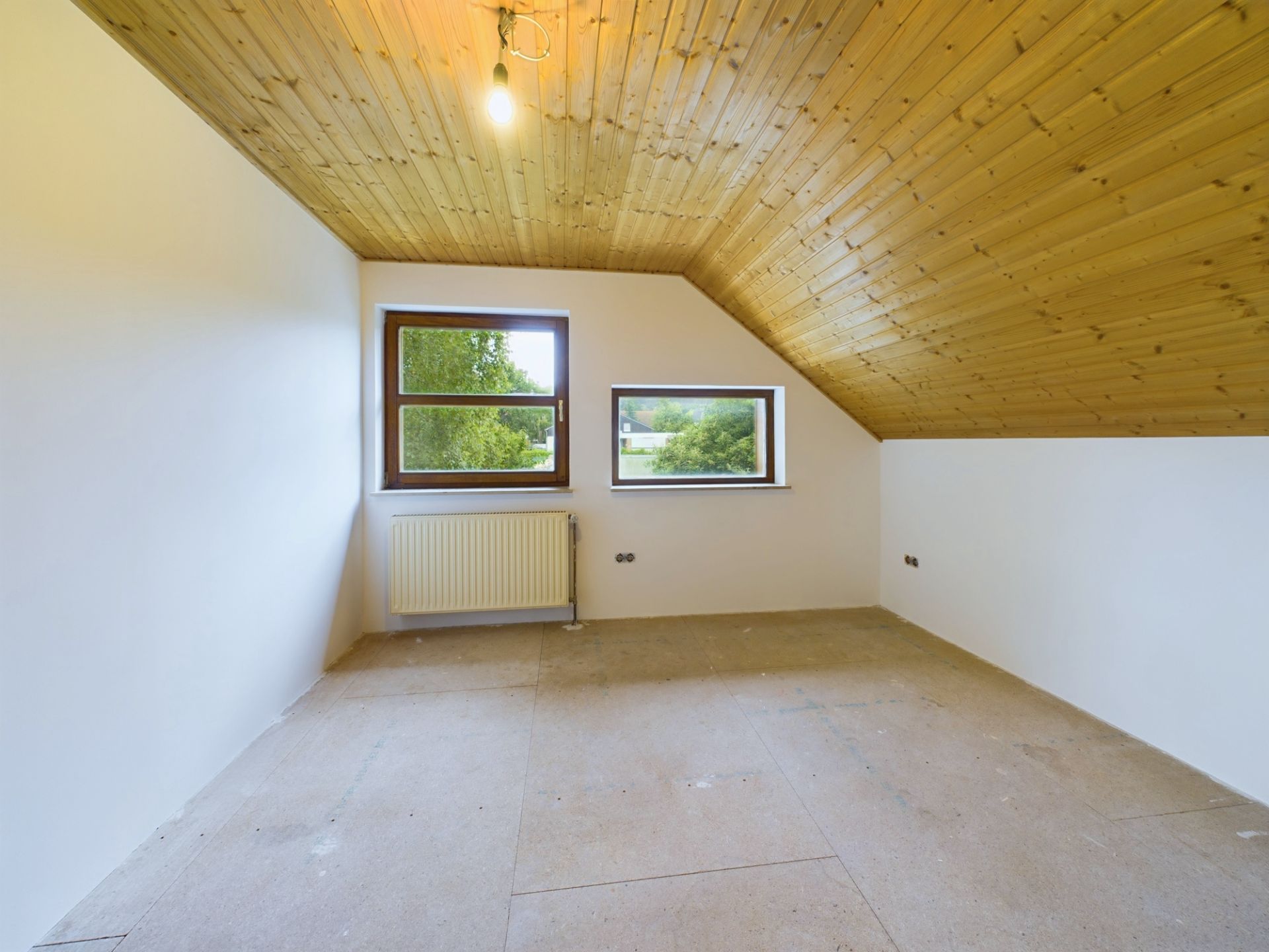 Leeres Zimmer mit schräger Holzdecke, zwei Fenstern und weiß gestrichenen Wänden.