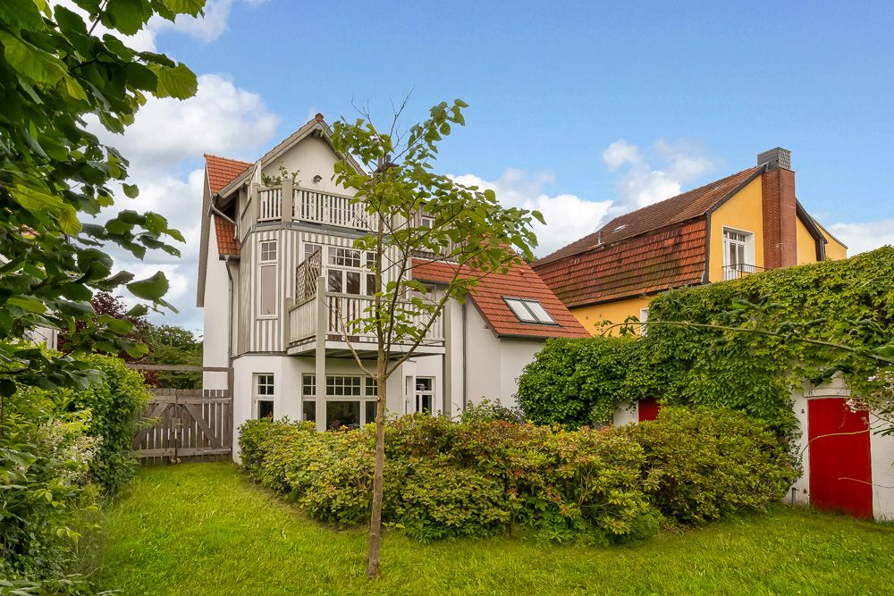 Zweistöckiges Wohnhaus mit Balkon und Garten, dicht umgeben von Büschen und einem Baum im Vordergrund.