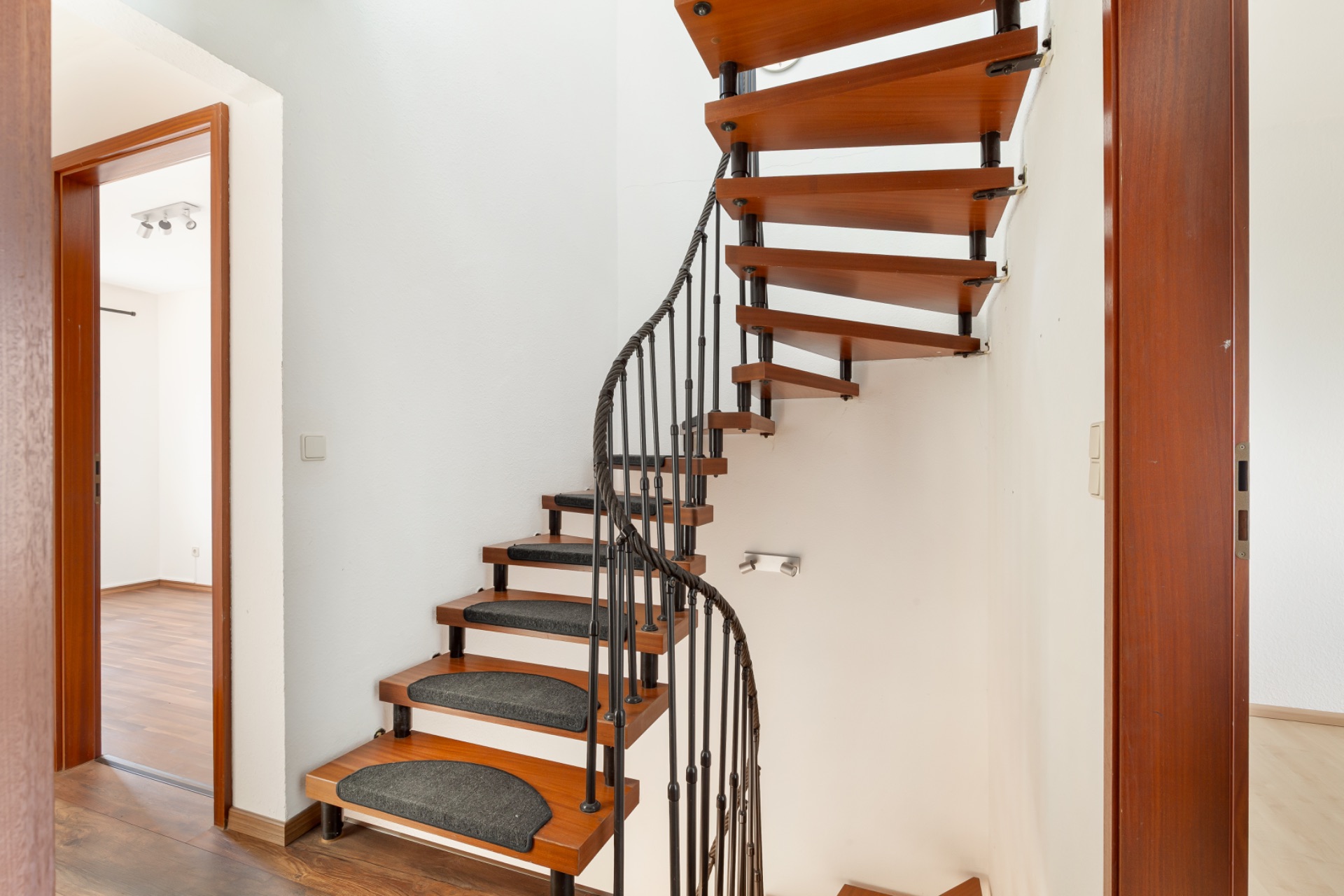 Holz-Wendeltreppe mit schwarzem Geländer in einem Flur mit Zugang zu zwei Zimmern.
