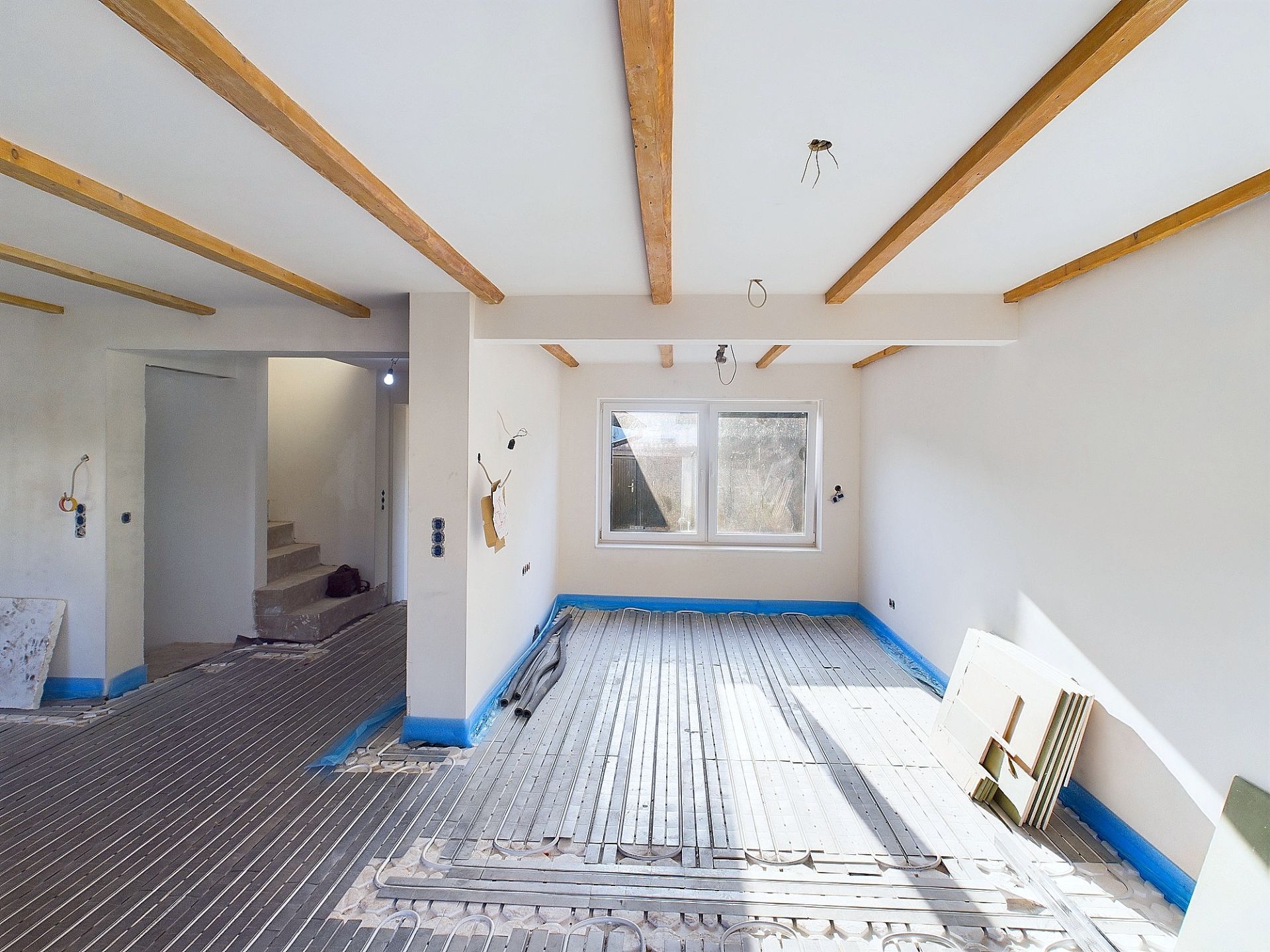 Heller Raum mit sichtbaren Holzbalken, großen Fenstern und unfertigem Boden in einem Haus.