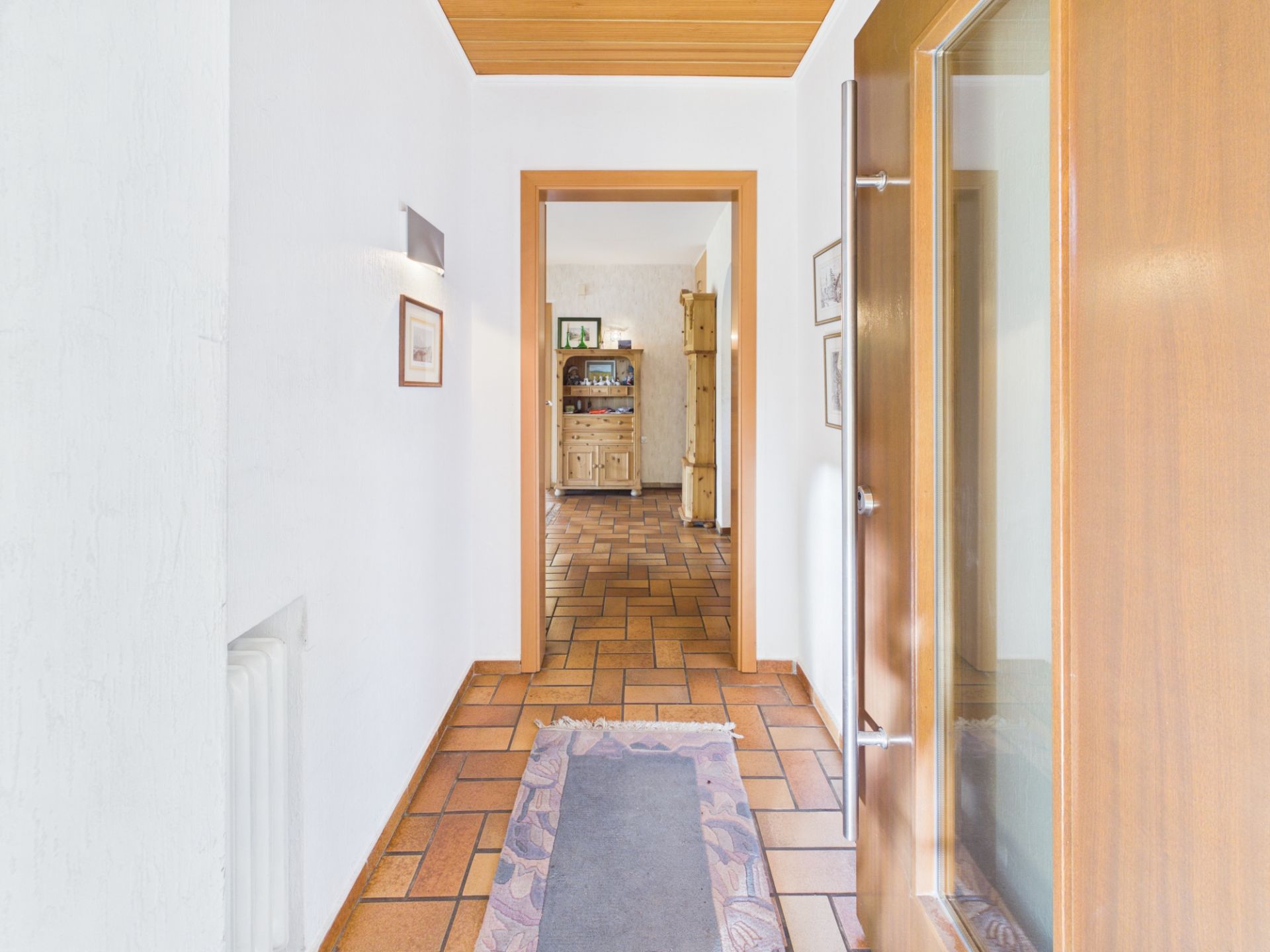 Heller Flur mit Fliesenboden, Teppichläufer und Blick in ein Wohnzimmer mit Holzschrank.