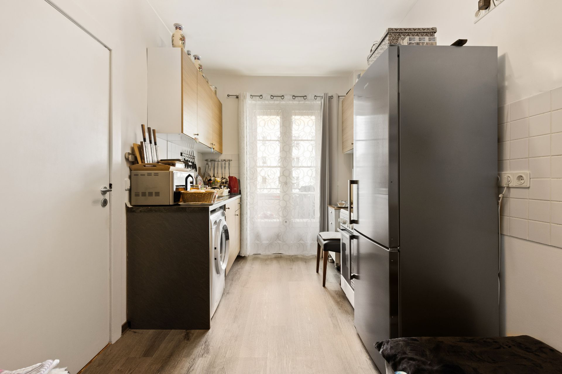Schmale Küche mit grauem Kühlschrank, Waschmaschine und Fenster mit Vorhang am Ende.