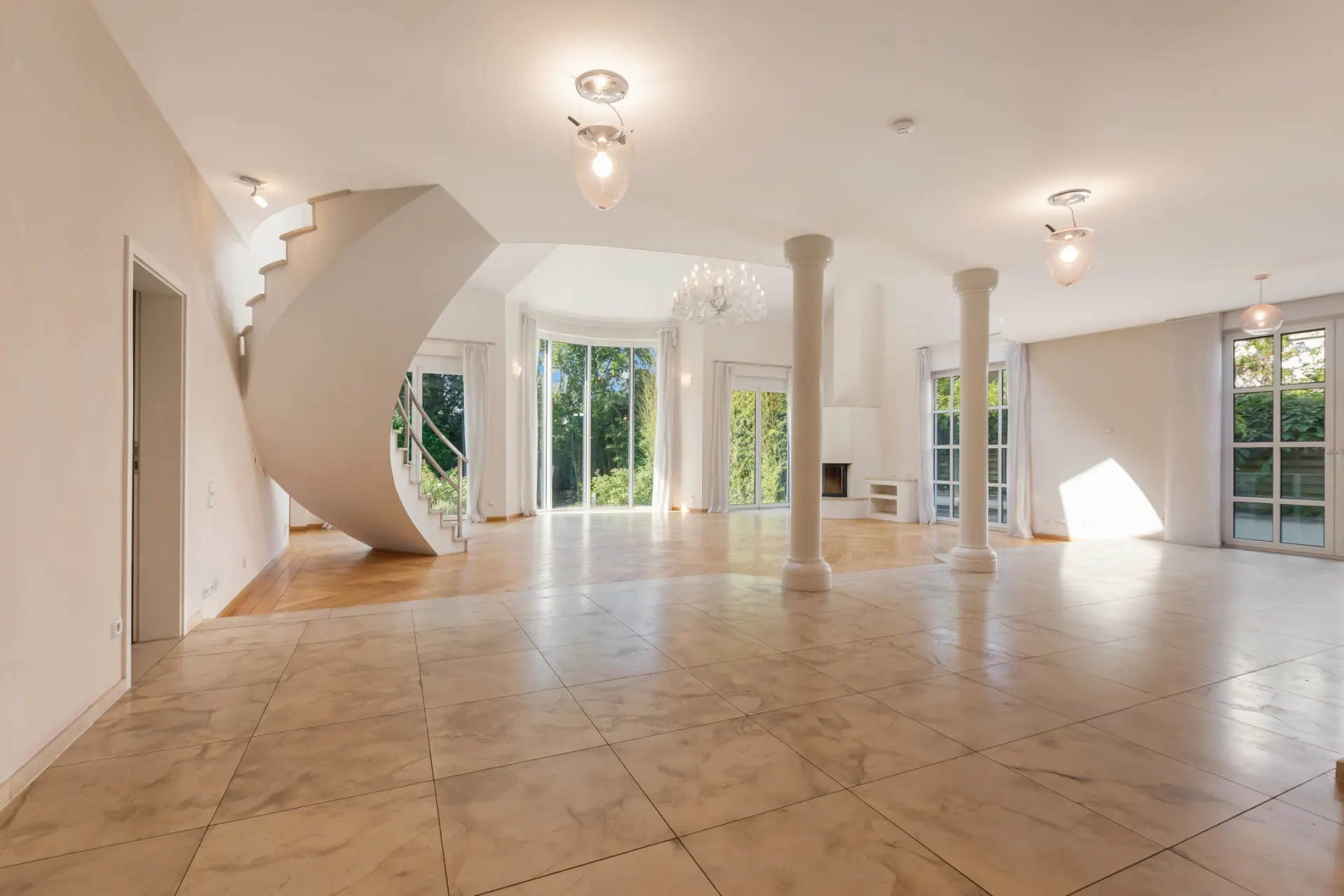 Großes, helles Wohnzimmer mit Marmorboden, Säulen, runder Treppe und großen Fenstern.