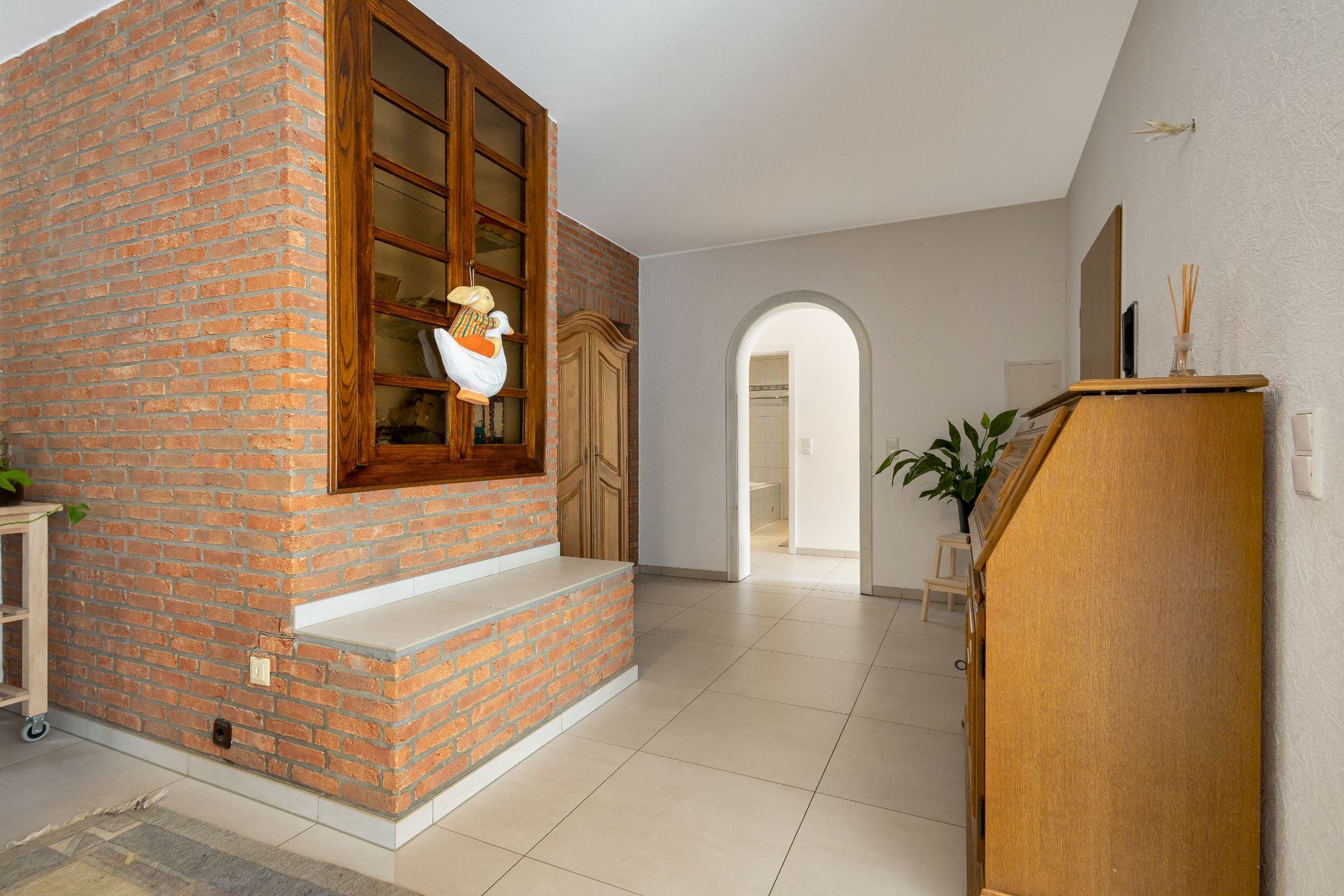 Heller Flur mit Backsteinwand, Holzdetails und Pflanzen, sichtbar sind Türen und ein Bogen.
