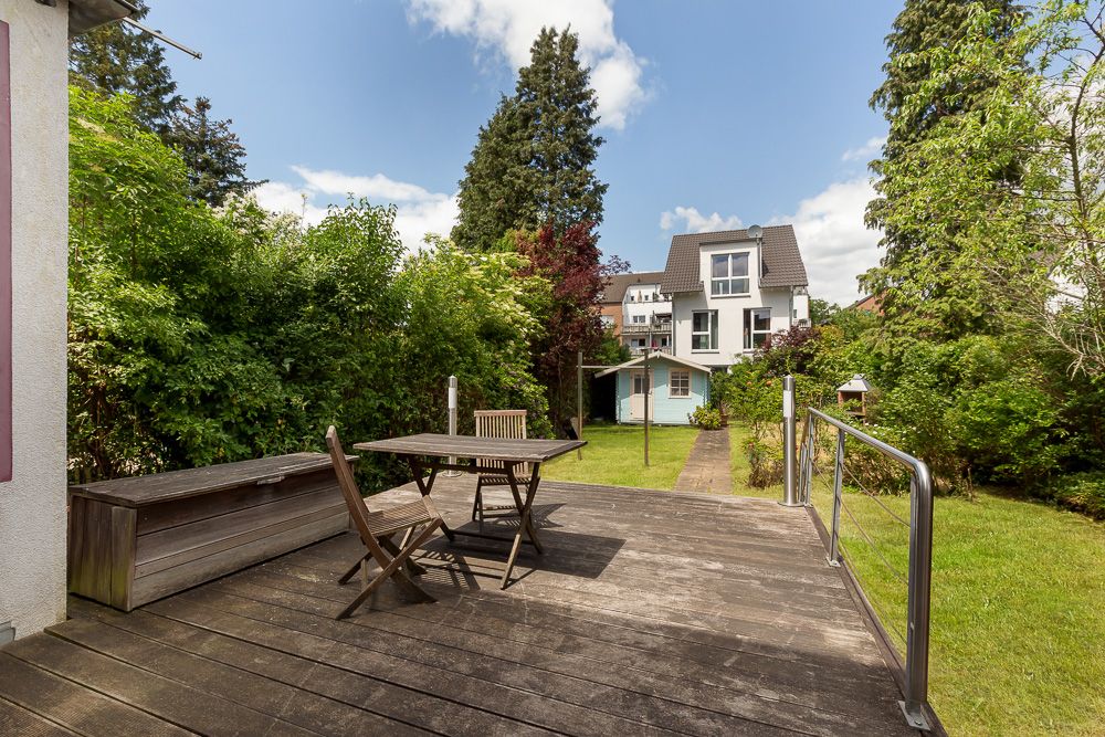 Holzterrasse mit Tisch und Stühlen im Garten, Blick auf ein Wohnhaus und ein Gartenhaus.