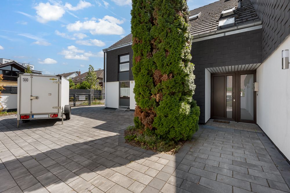Gepflasterter Hof mit Anhänger und großem Baum vor modernem Wohnhaus.