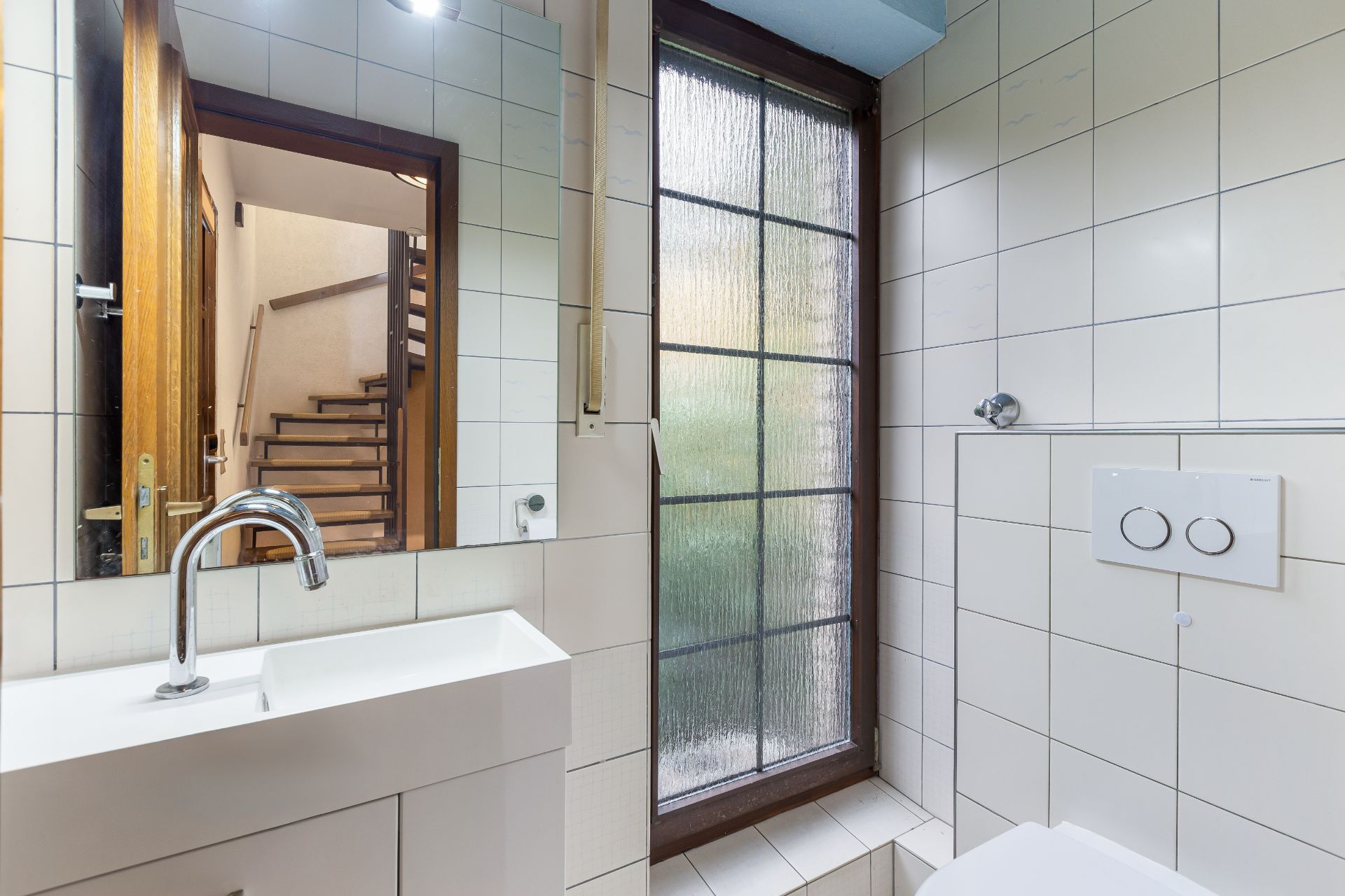 Kleines Badezimmer mit Waschbecken, Spiegel, Fenster und weiß gefliester Wand.