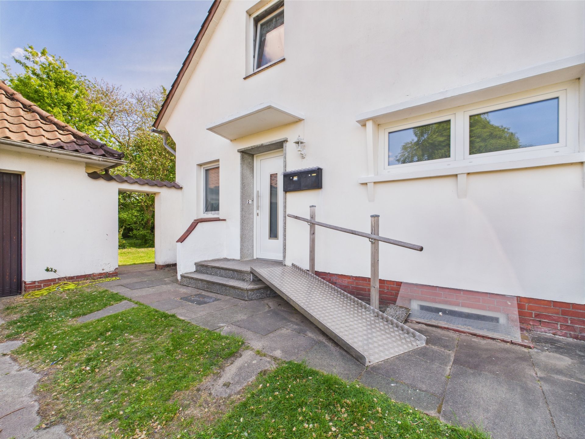 Eingang eines Wohnhauses mit Rampe und Treppe, umgeben von Grünfläche.
