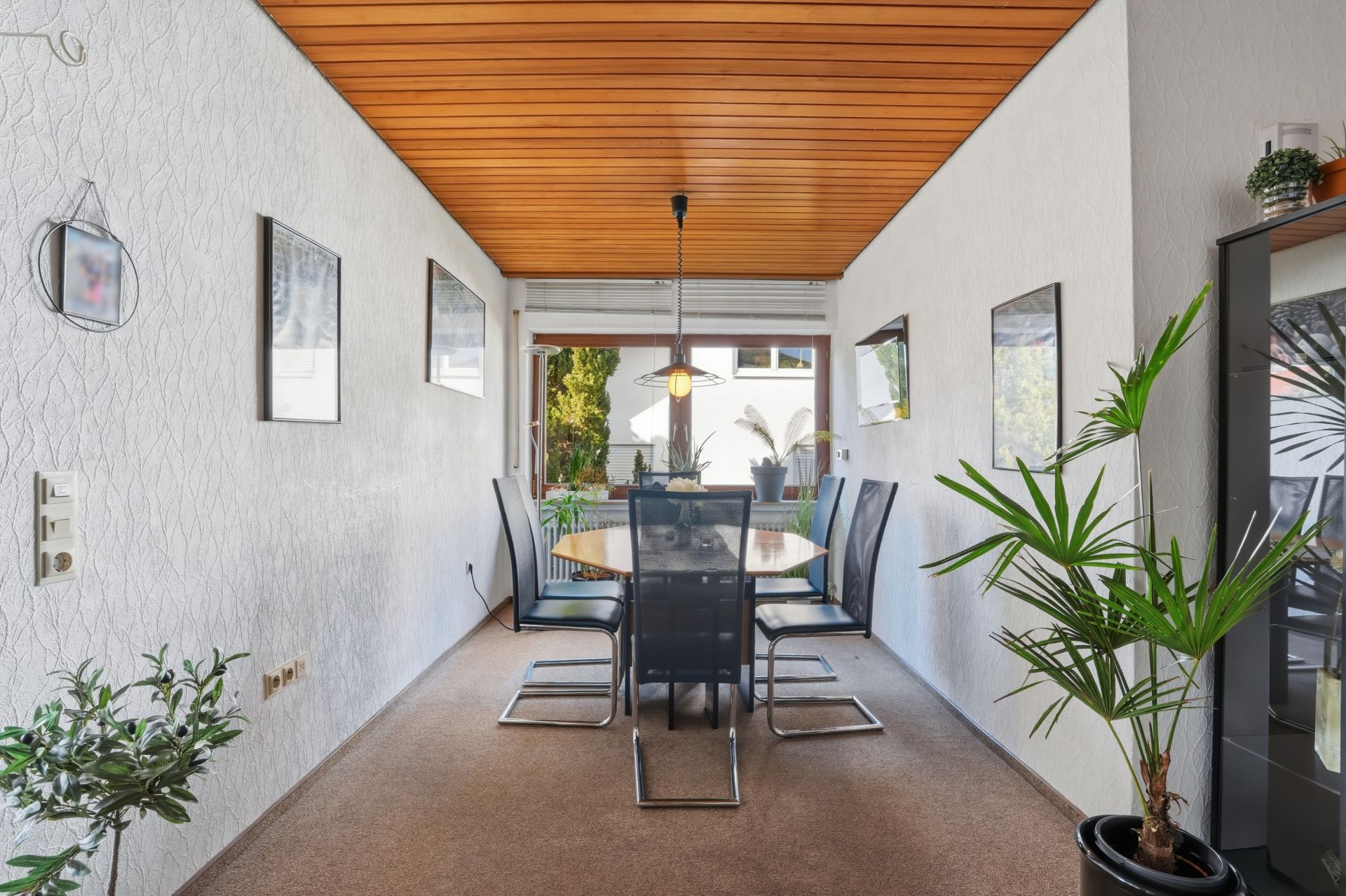 Esszimmer mit rundem Tisch, sechs Stühlen, Pflanzen, Bildern an den Wänden und Fensterblick ins Grüne.