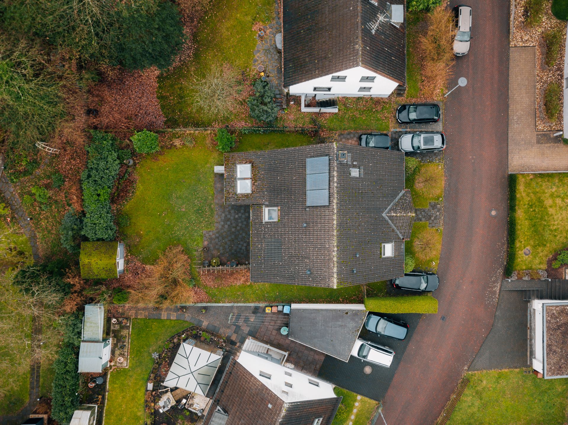 Luftaufnahme eines Hauses mit Garten, geparkten Autos und angrenzender Straße.