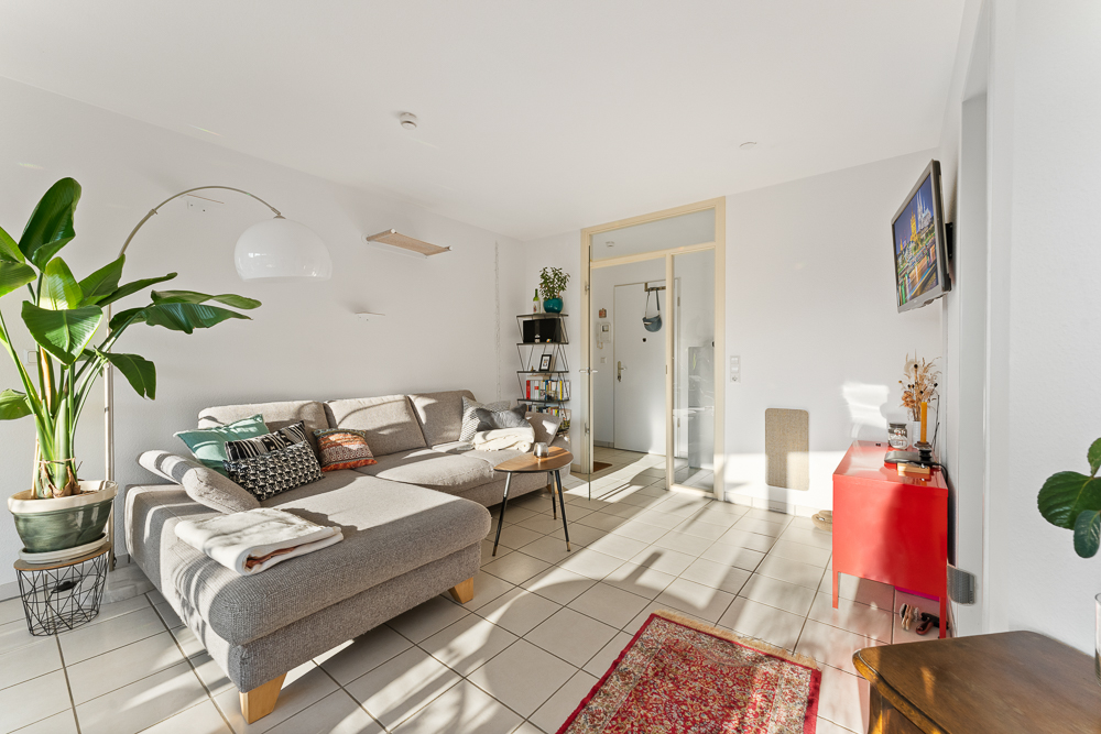 Helles Wohnzimmer mit grauem Ecksofa, Pflanzen, kleinem Tisch und rotem Sideboard.