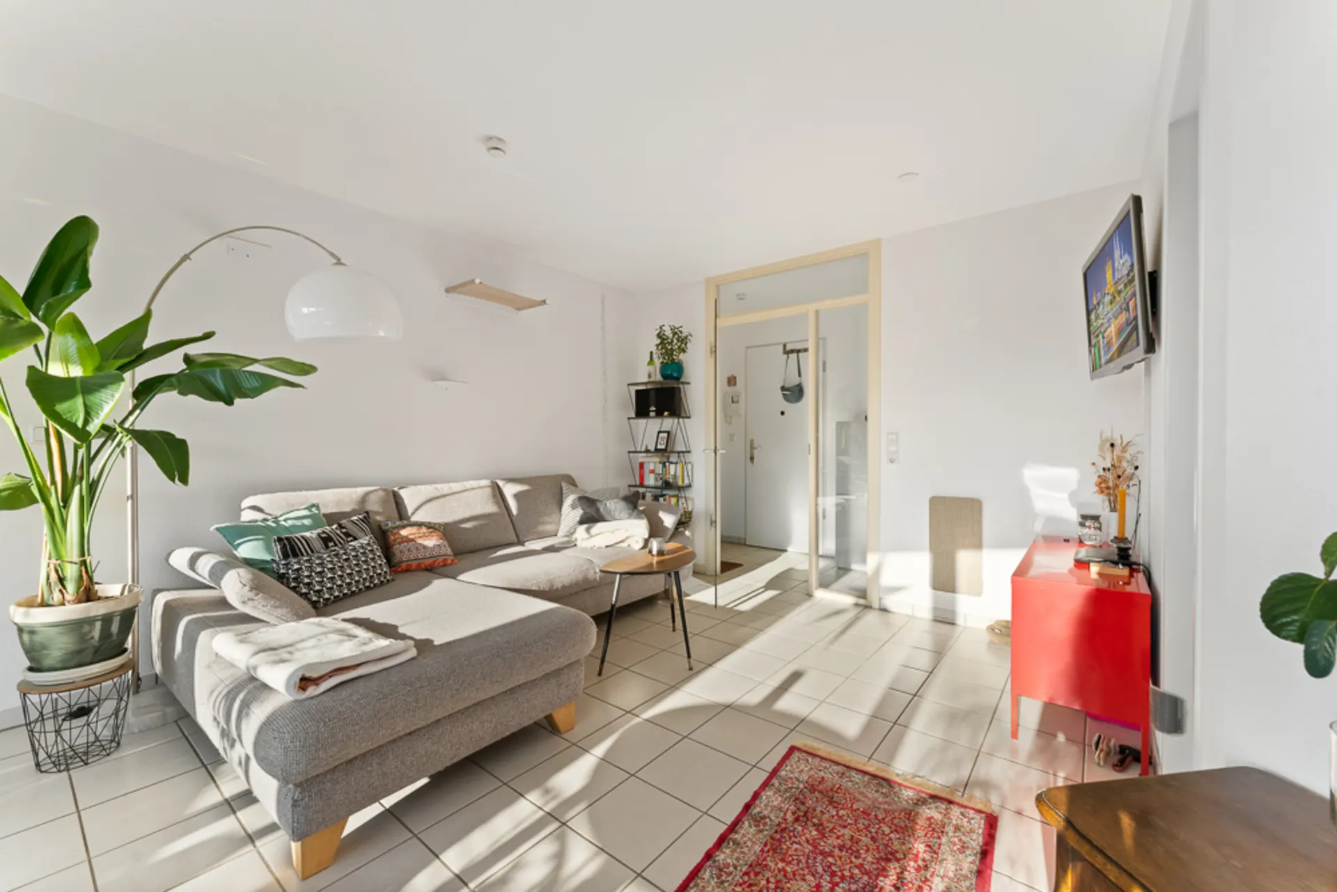 Helles Wohnzimmer mit grauem Ecksofa, Pflanzen, kleinem Tisch und rotem Sideboard.