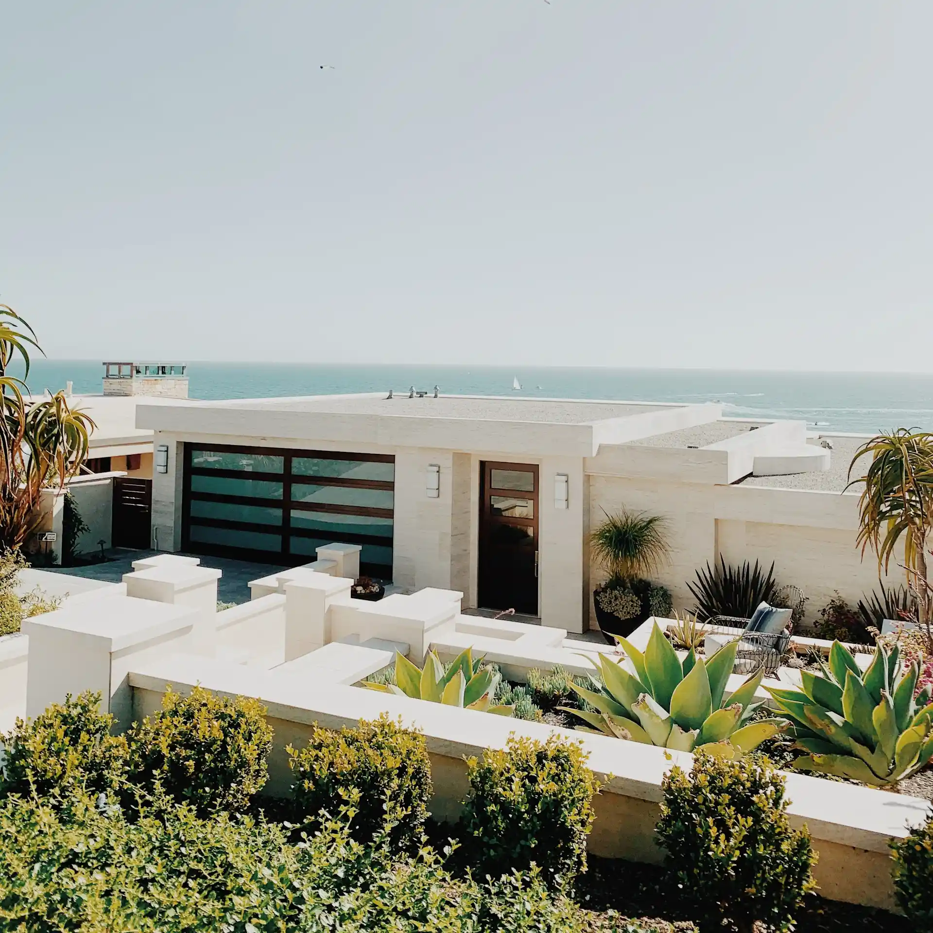 Modernes Haus mit Flachdach und großem Fenster, Blick auf das Meer, umgeben von Pflanzen.