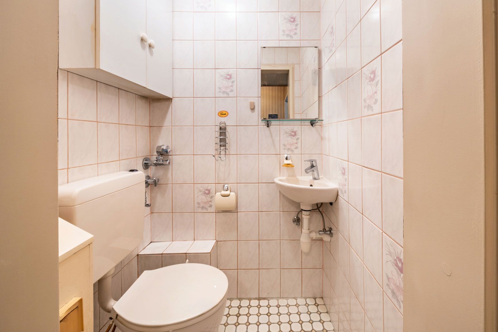 Kleines Badezimmer mit Toilette, Waschbecken, Spiegel und hellen Fliesen mit Blumenmuster.