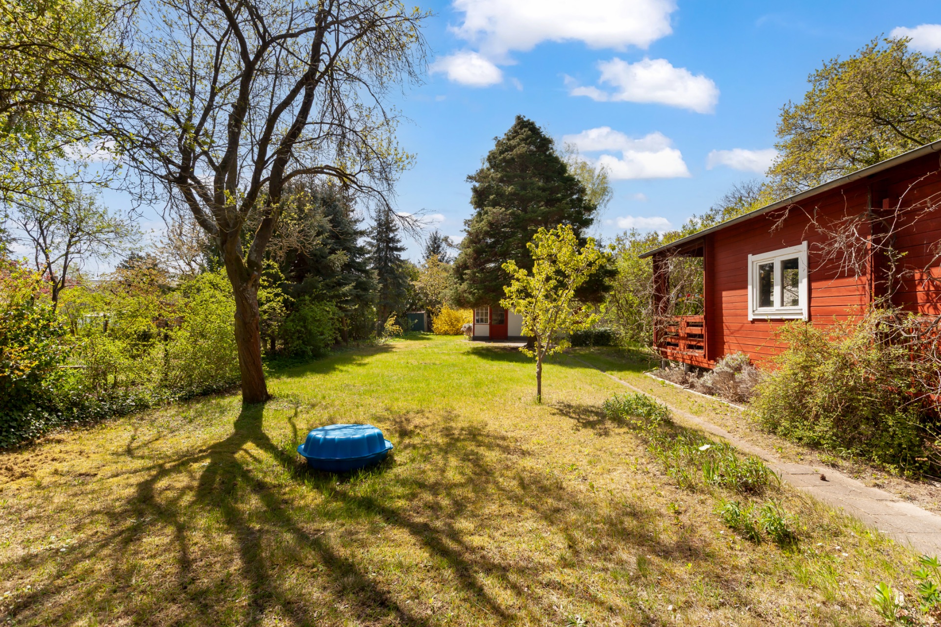 Garten mit Rasen, Bäumen und einem roten Holzhaus rechts.