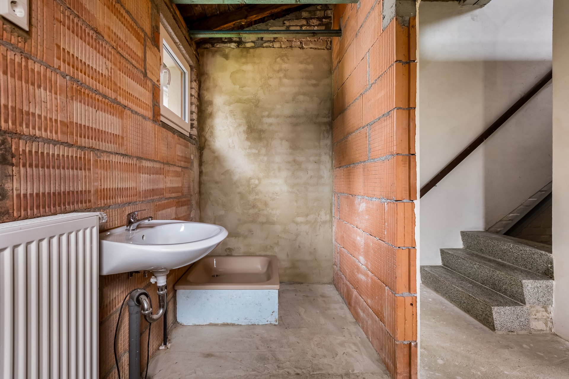 Unfertiges Bad mit Waschbecken, Heizung und Duschwanne neben einer Betontreppe.