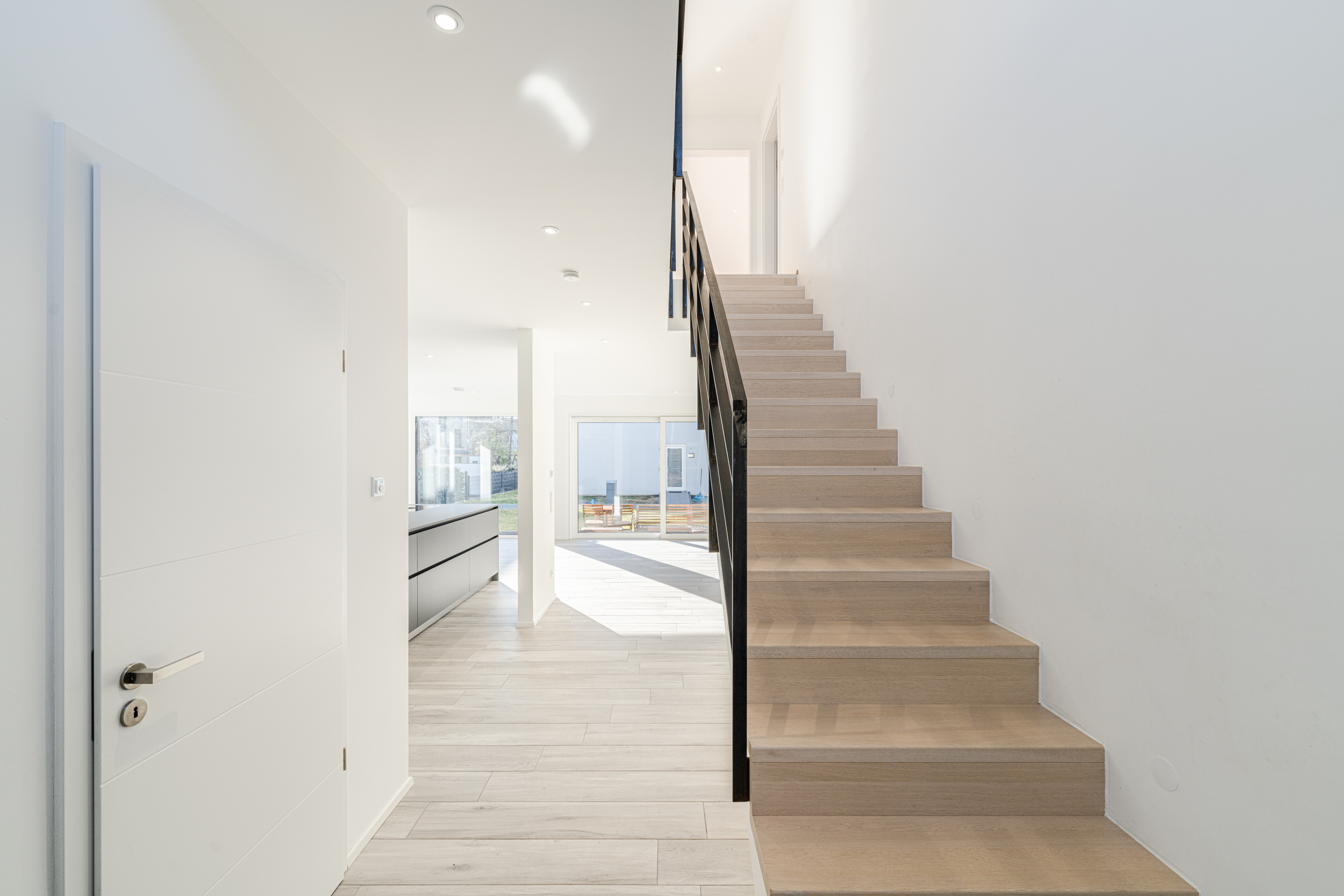 Helle Diele mit Holztreppe, weißer Tür und Blick auf Küche und Garten.