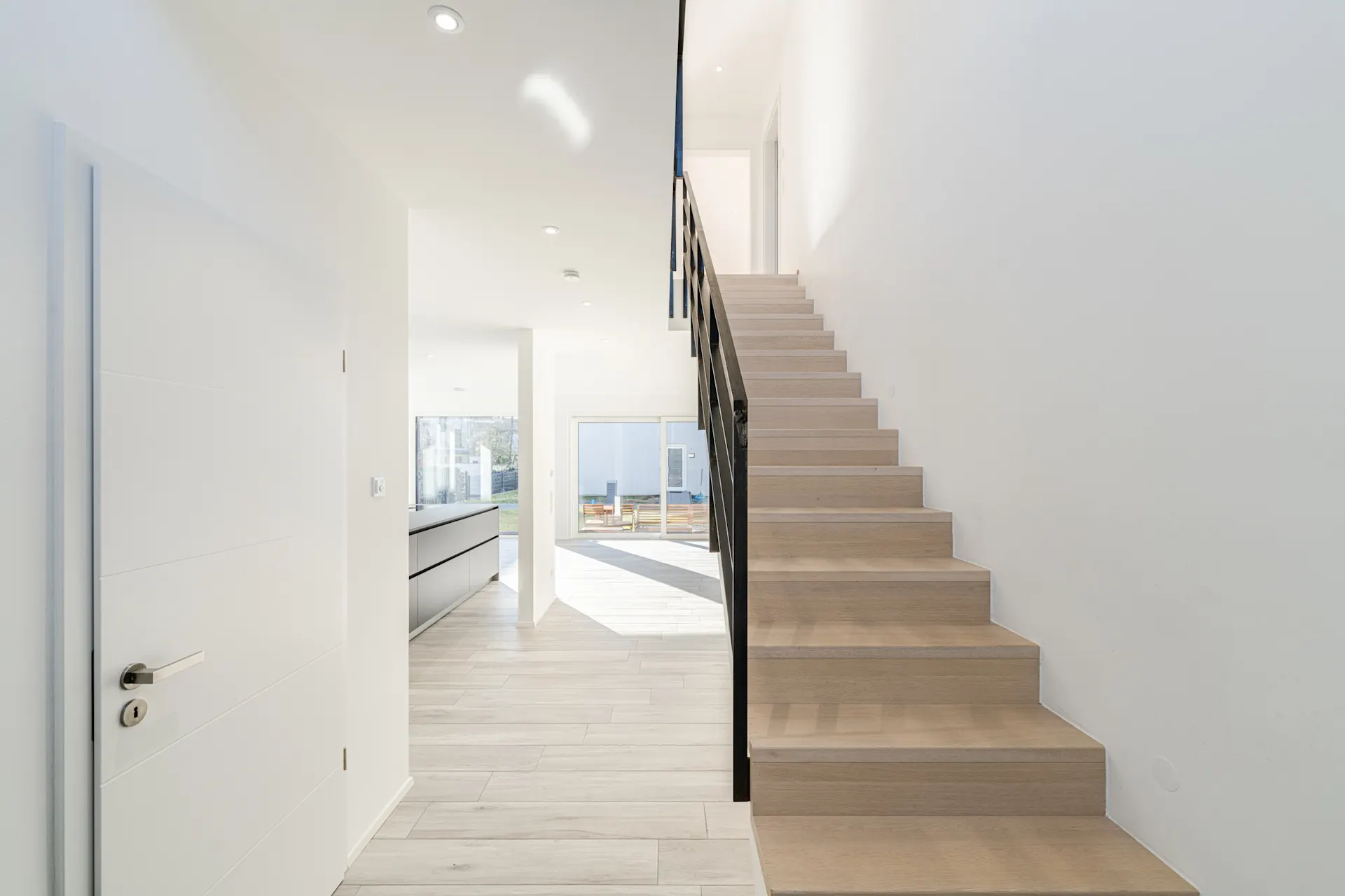Helle Diele mit Holztreppe, weißer Tür und Blick auf Küche und Garten.