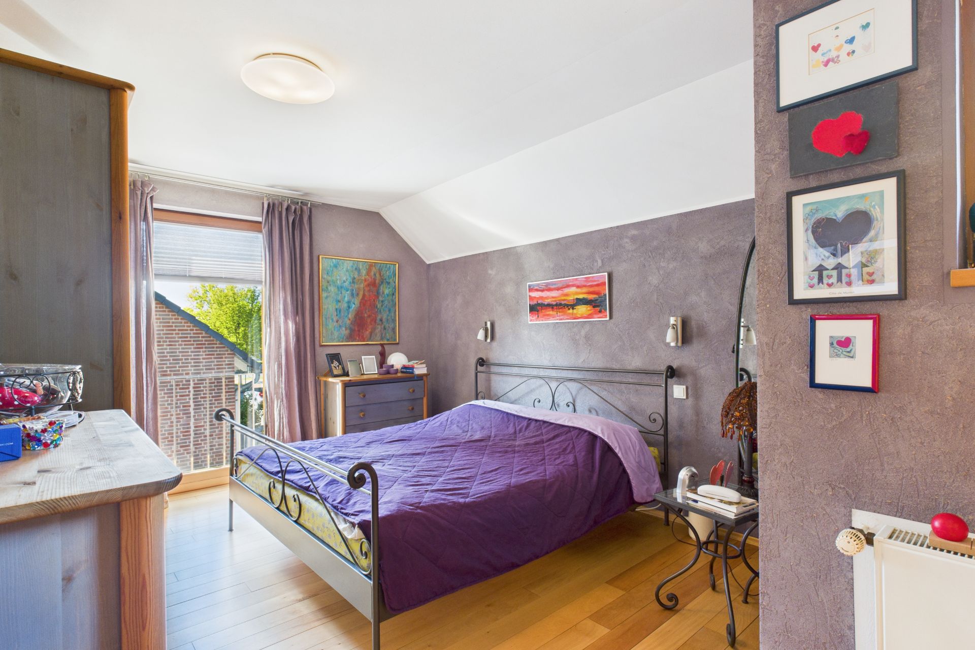 Schlafzimmer mit Metallbett, lila Decke, Holzboden und Bildern an den Wänden.