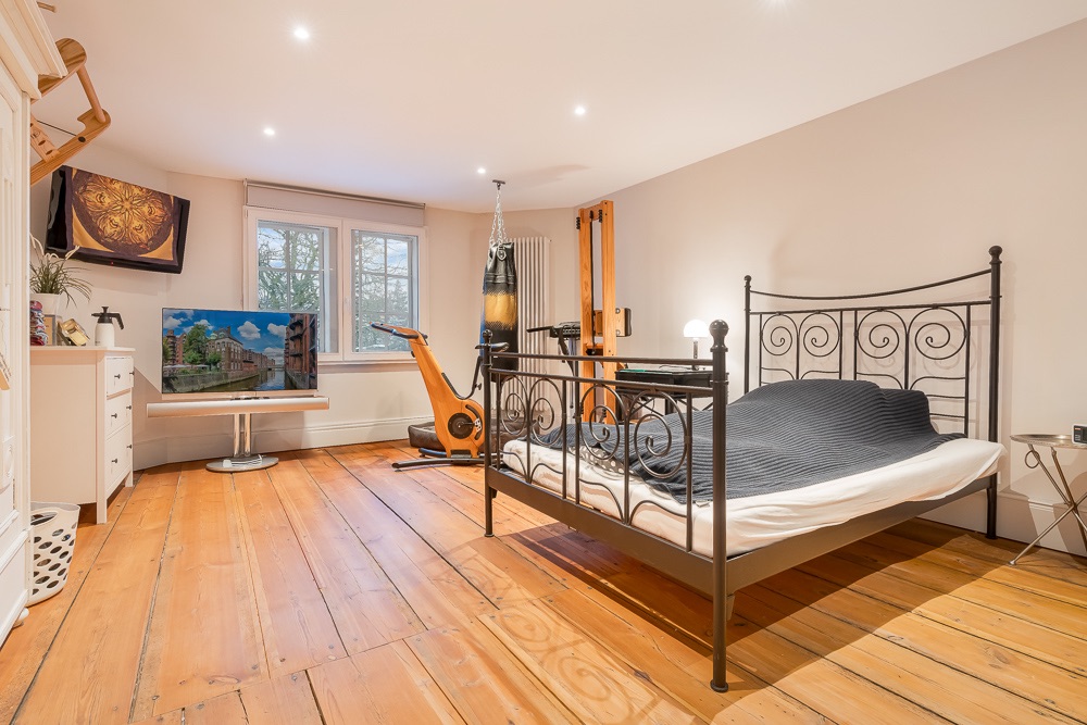 Helles Schlafzimmer mit Metallbett, Holzboden, Fernseher, Fitnessgerät und zwei Fenstern.