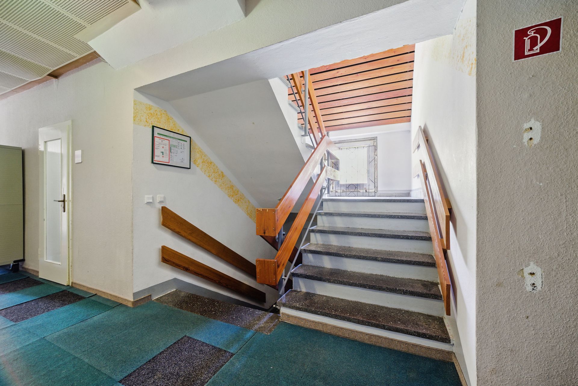 Treppe mit Holzgeländer in einem Wohngebäude, grün gemusterter Boden, Tür links.